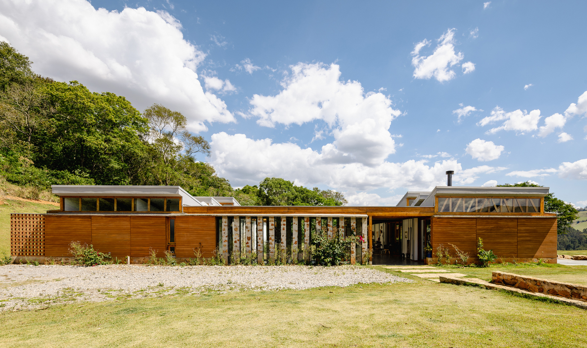 Low timber-clad house with clerestory windows and a planted screen of stone piers, seen across gravel under cumulus clouds