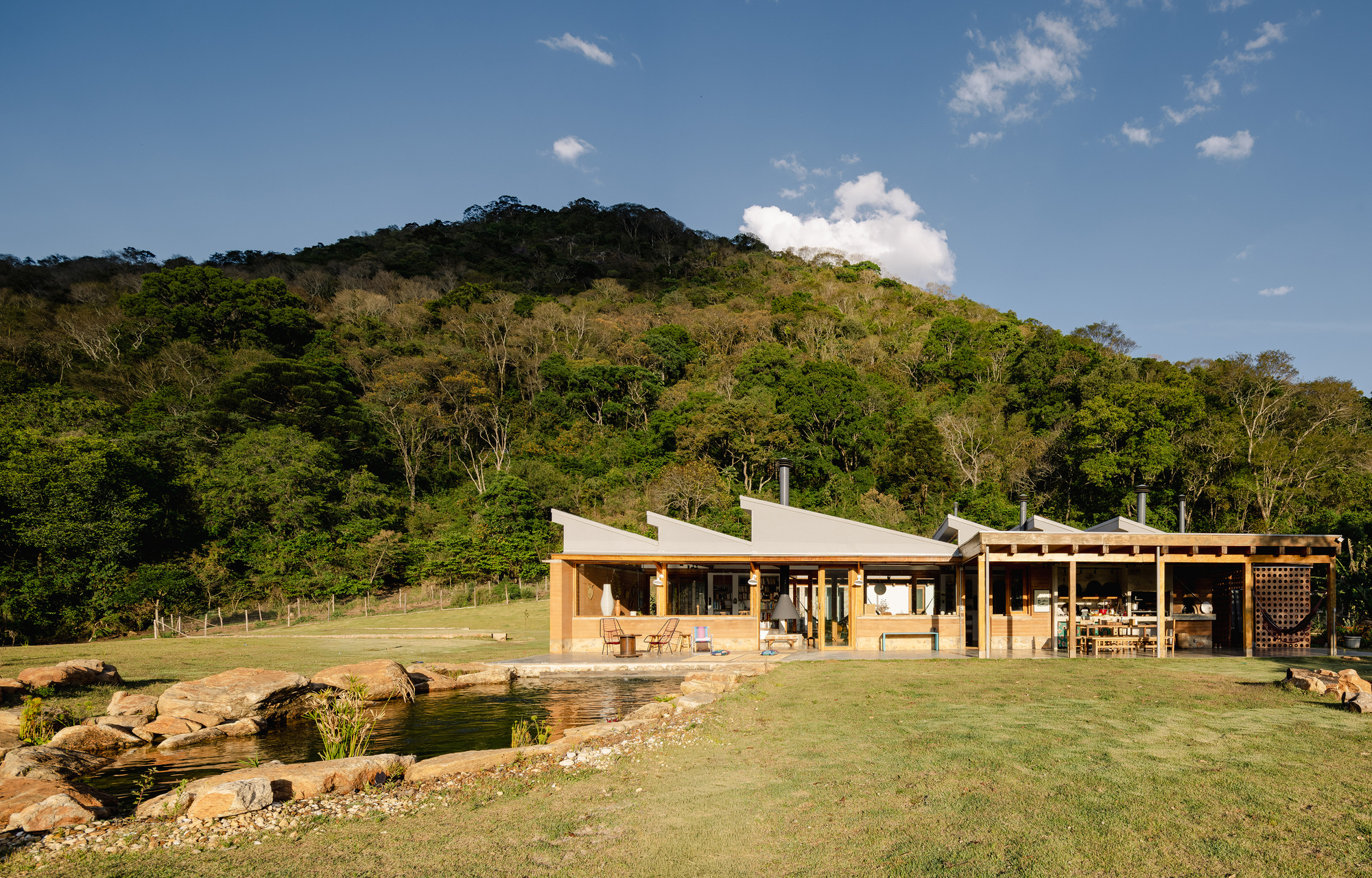Rear facade with sawtooth roof and open porch beside a stone-edged pond, with a forested hill rising behind in afternoon light