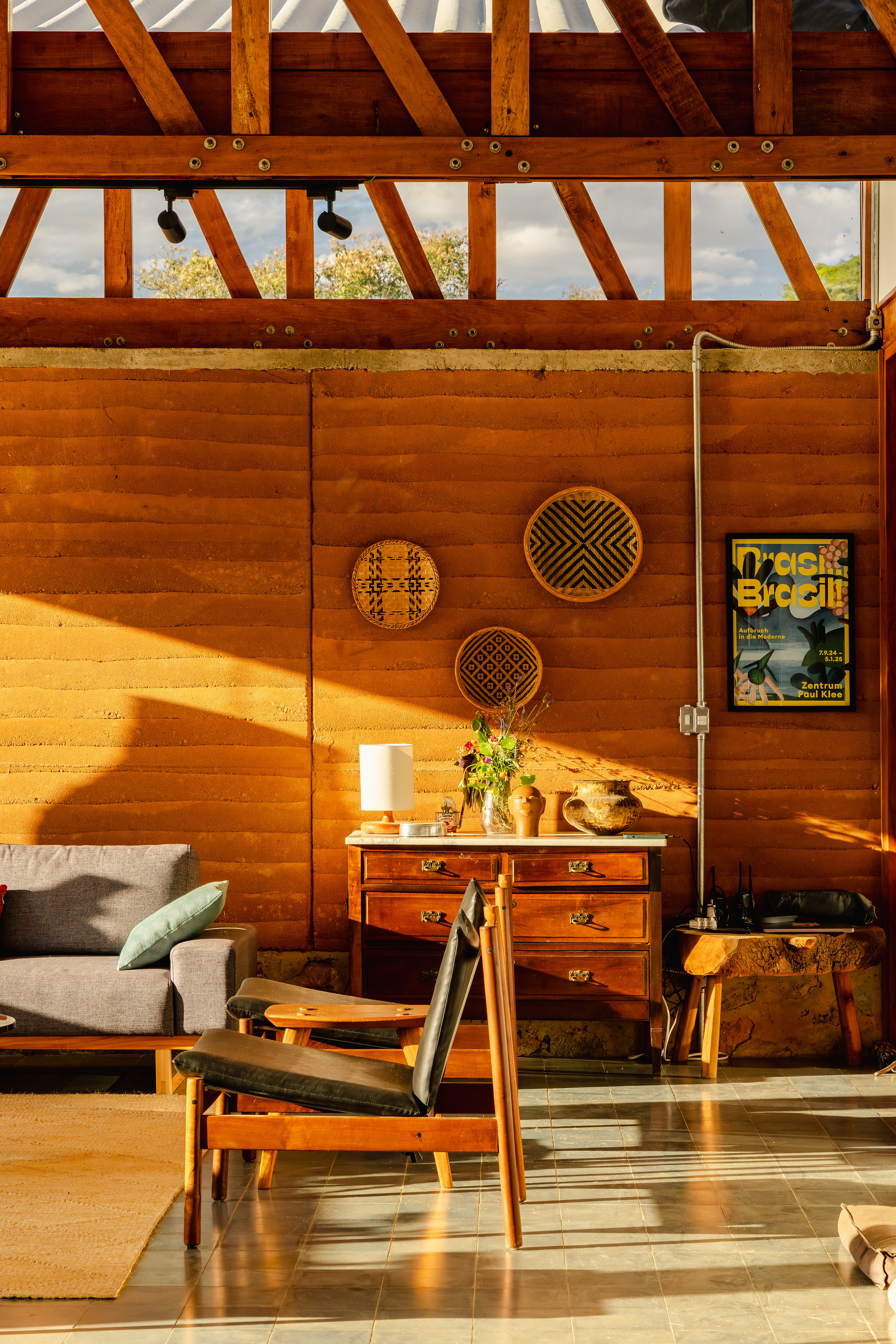 Rammed earth living room wall with a wooden dresser, woven wall discs and leather lounge chair in low golden sunlight