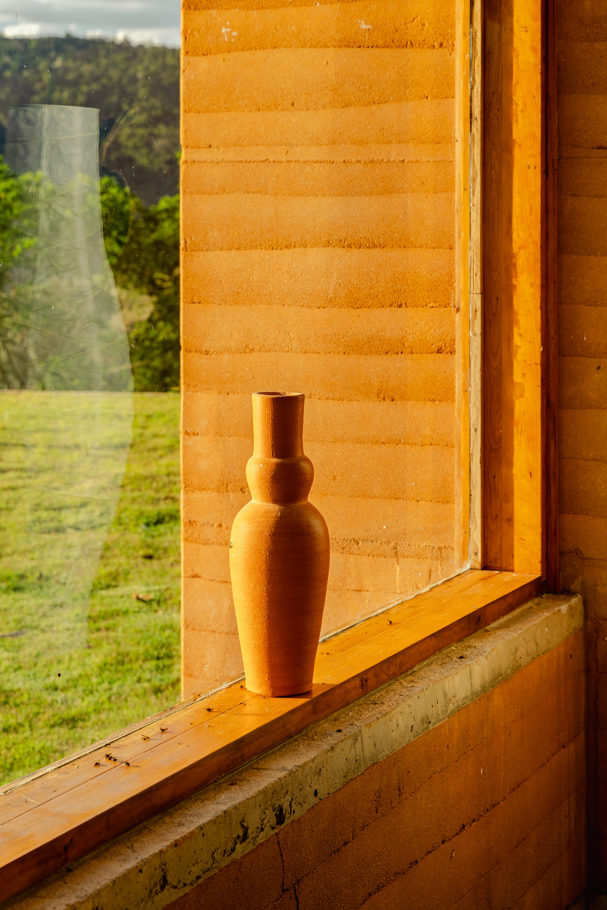 Terracotta vase on a timber window sill against a rammed earth wall, with a green lawn beyond in afternoon light