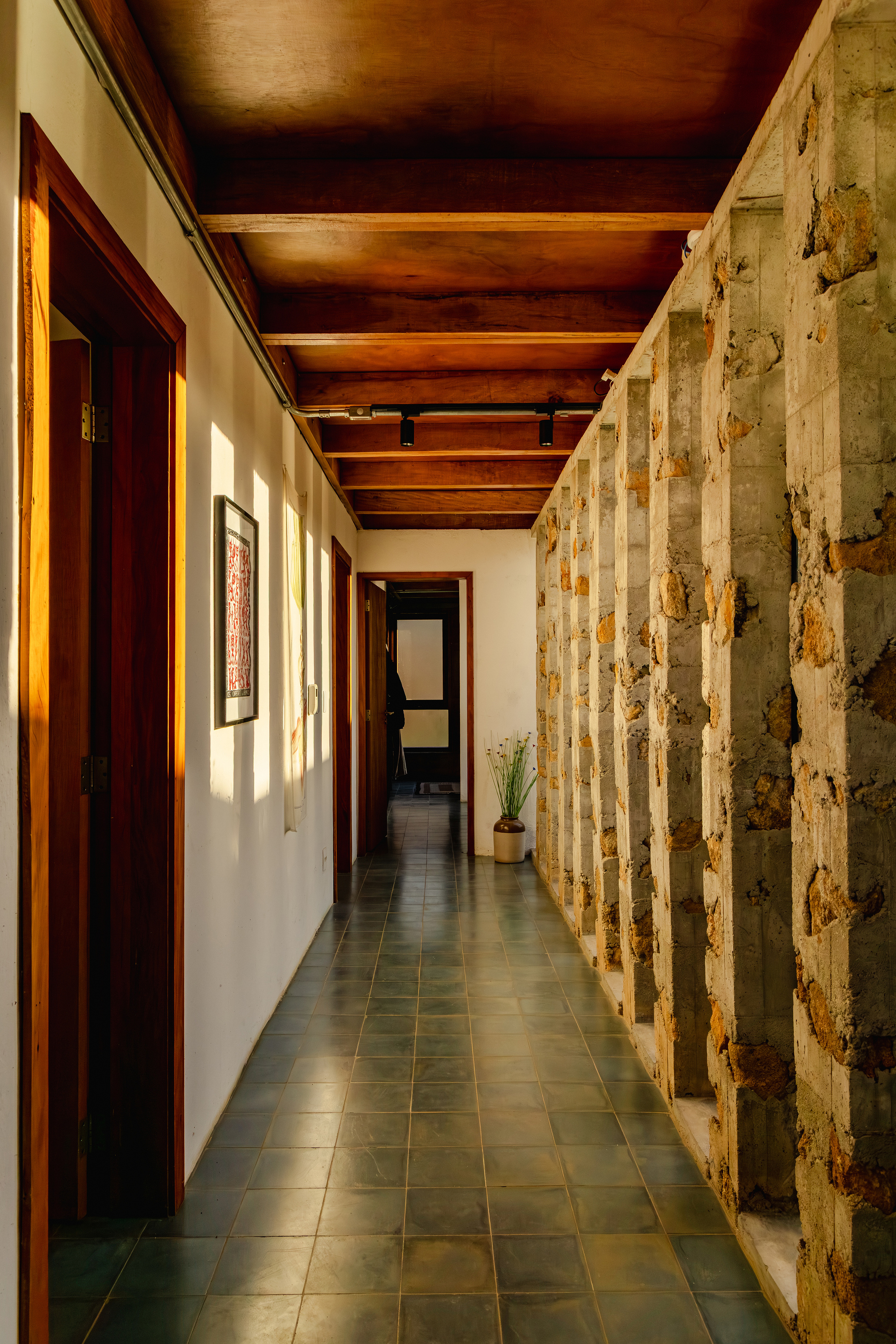 Corridor with a timber beam ceiling, slate floor tiles and a stone and concrete column wall, lit by low sun