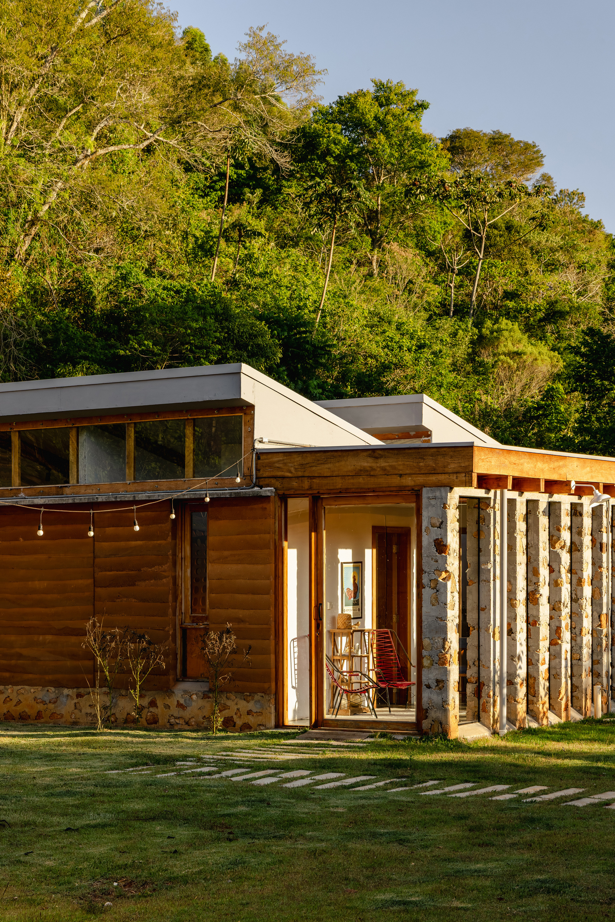 Exterior of a timber-clad pavilion with a row of stone columns and string lights, set against a forested hillside