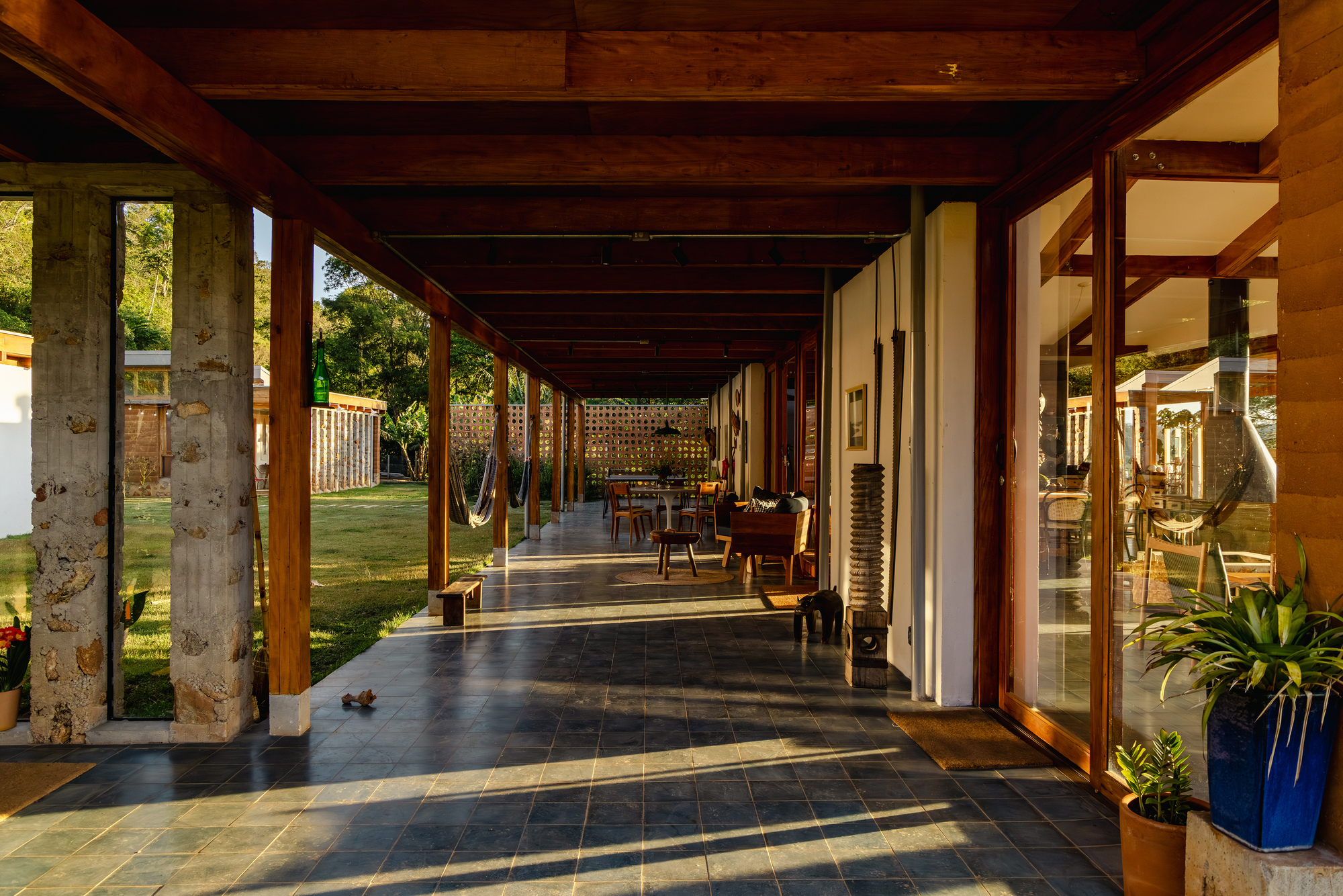 Covered veranda with stone-faced columns, timber posts and slate paving, striped by late afternoon shadows and a hammock in the distance