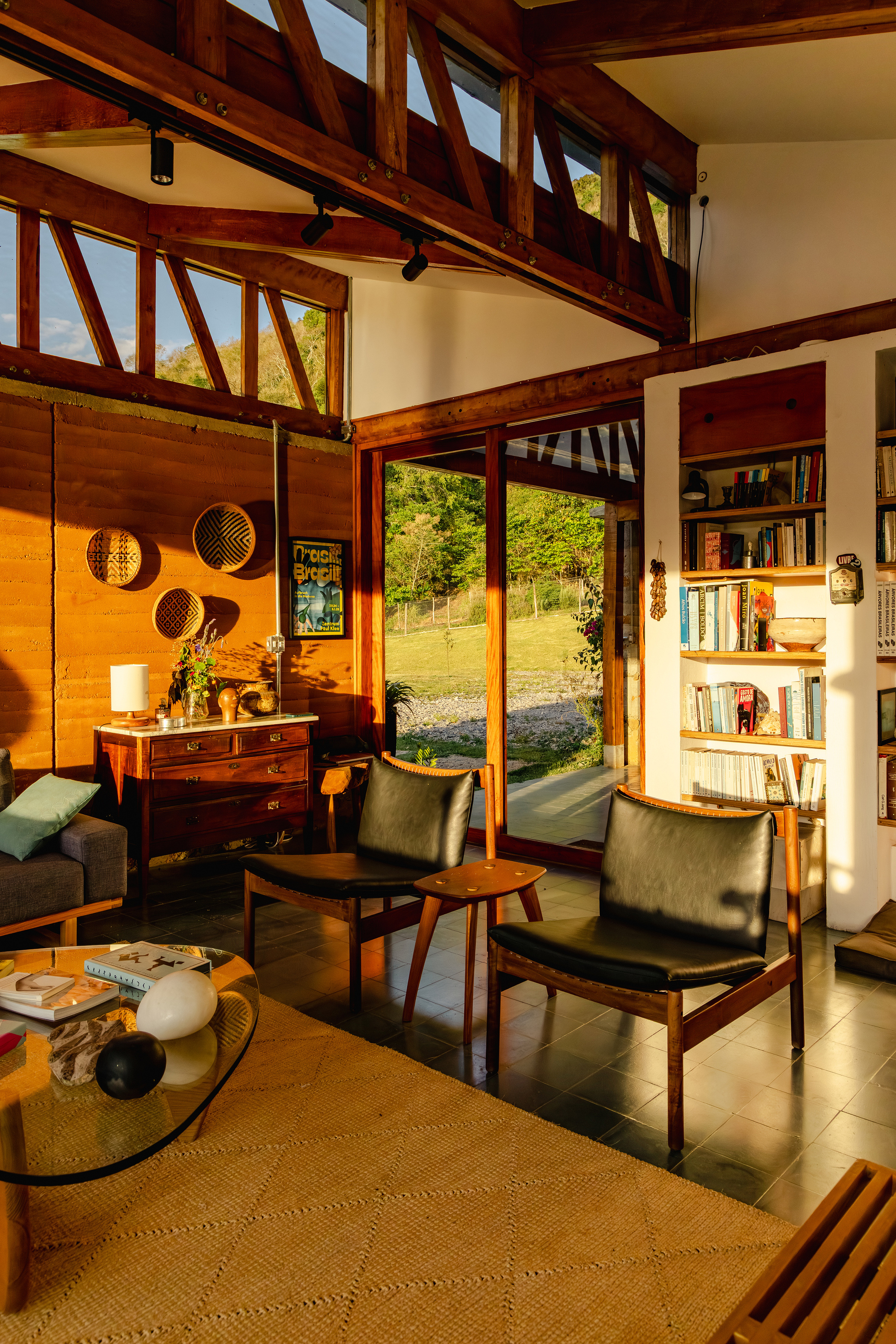 Double-height living room with exposed timber trusses, built-in bookshelves and leather armchairs facing a glass door in warm evening sun