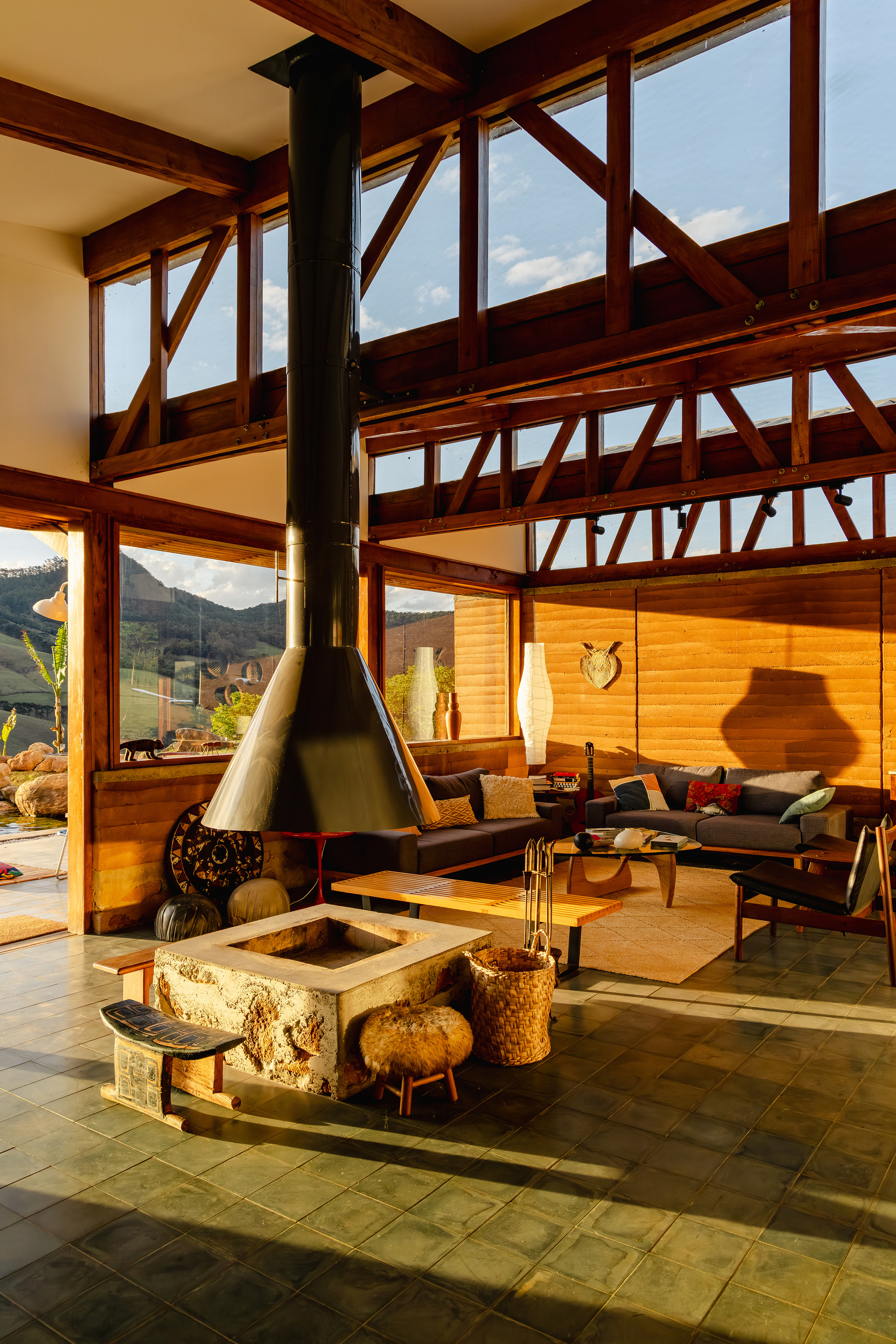 Double-height living room with a black conical hanging fireplace, stone fire pit and timber trusses in low afternoon sun