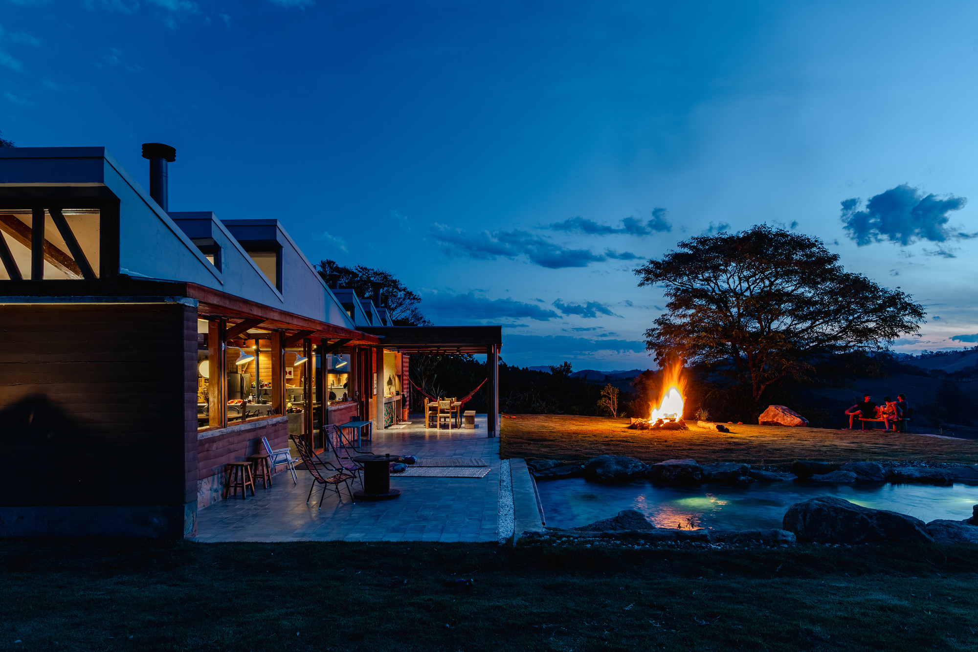 Low house with a covered terrace, a pond edged with boulders and a bonfire burning under a dusk sky