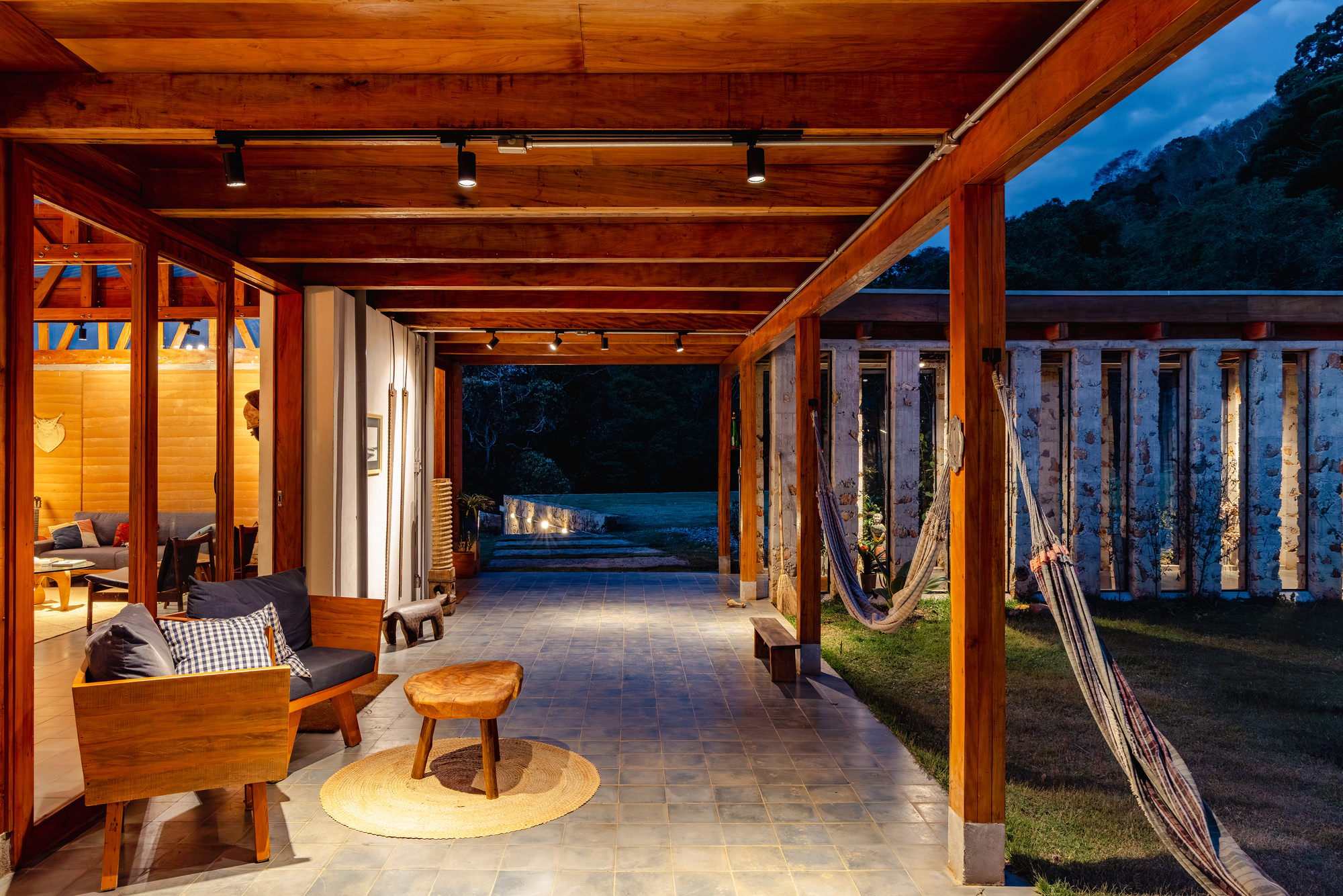 Timber-framed gallery walkway with track lights, a stone wall with narrow openings and hammocks hanging at blue hour