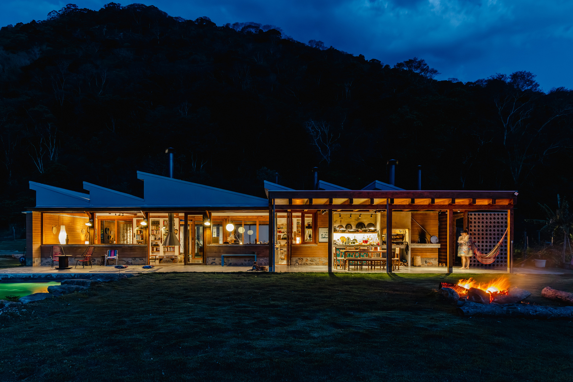 Long lit facade with sloped roofs and open kitchen pavilion, a bonfire in the foreground and a dark forested hill behind