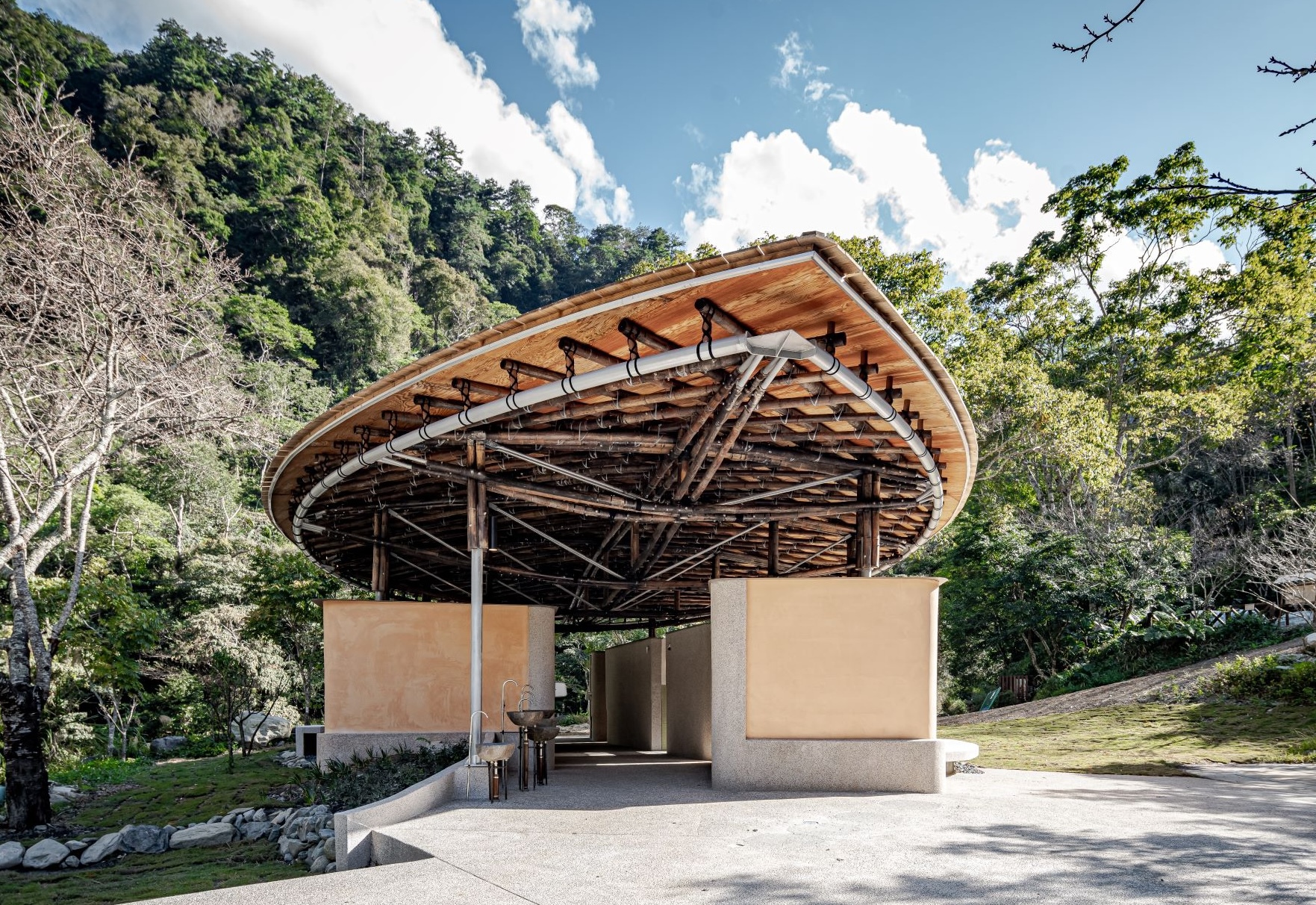 Round bamboo-framed canopy roof over ochre rammed earth walls, set against forested slopes and drifting clouds