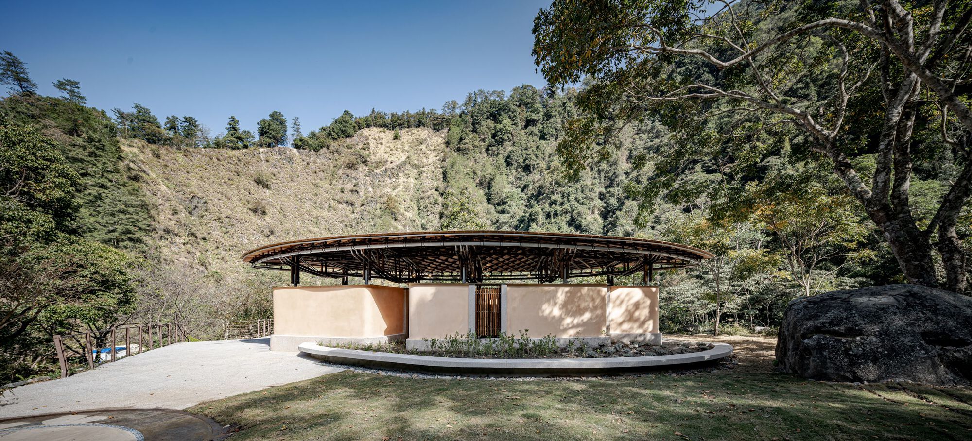 Low side view of the disc-shaped roof above ochre walls and a curved planter, with a boulder in the foreground