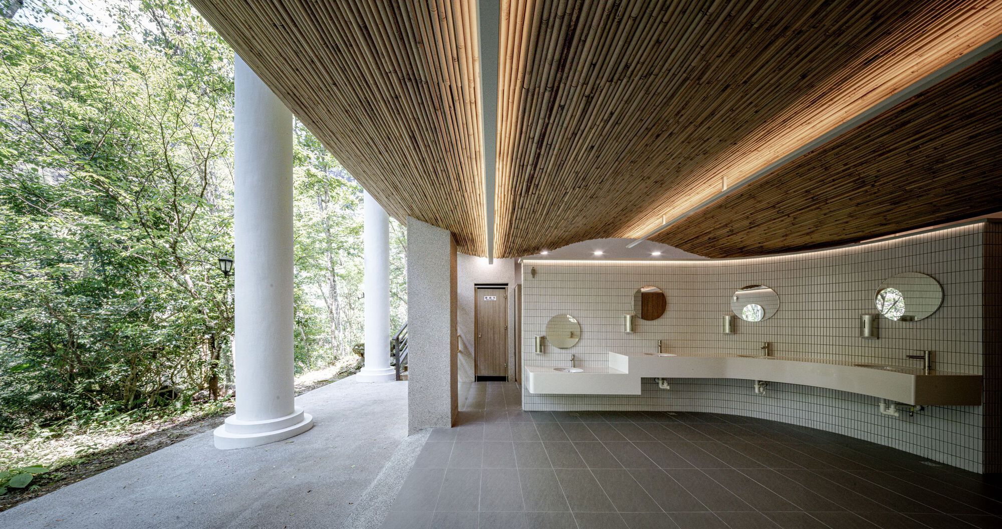 Restroom interior with a curved white tile wall, round mirrors and a slatted bamboo ceiling, white columns open to the forest