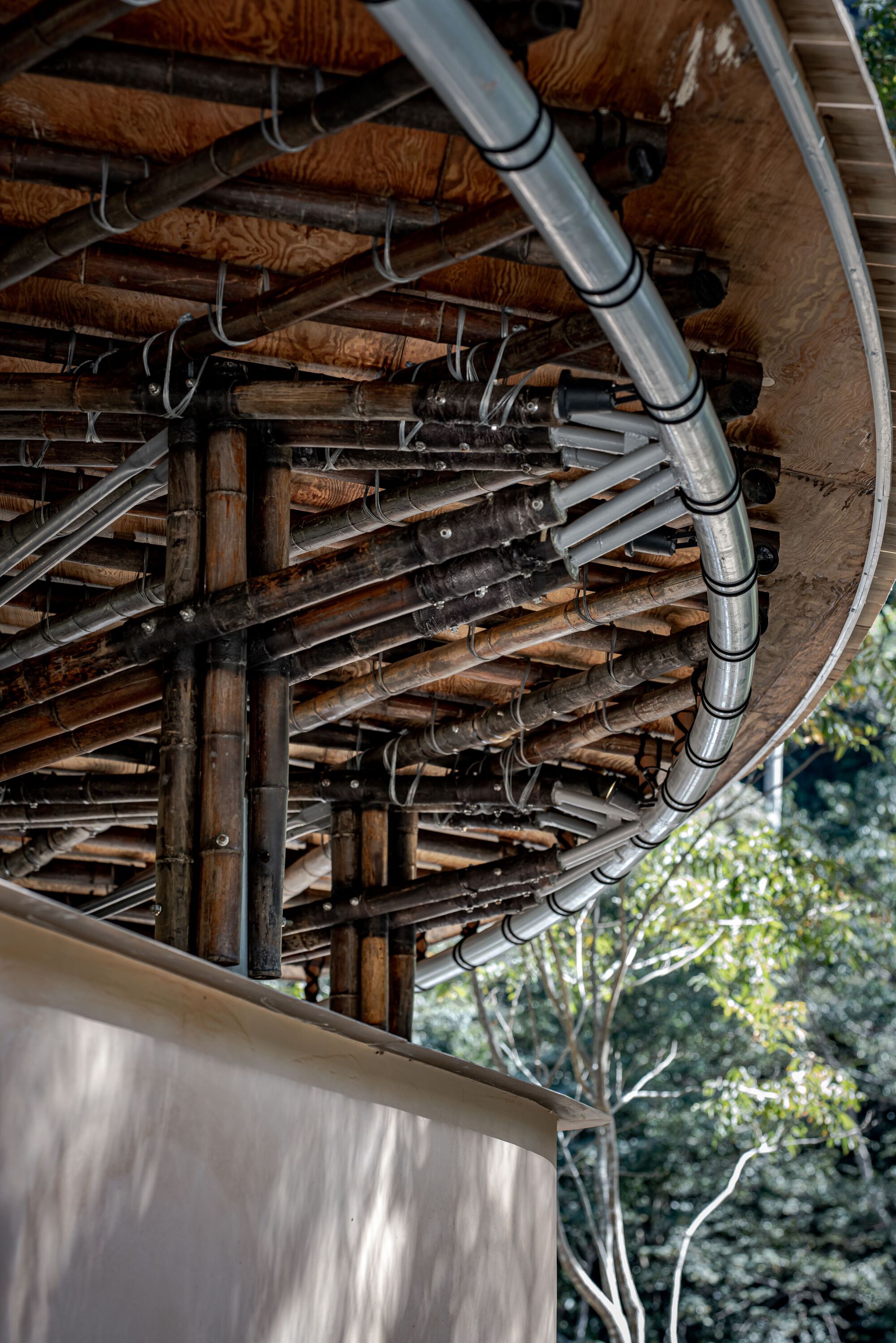 Underside of the curved roof showing bamboo poles, metal conduit and plywood decking above a pale earth wall in dappled light