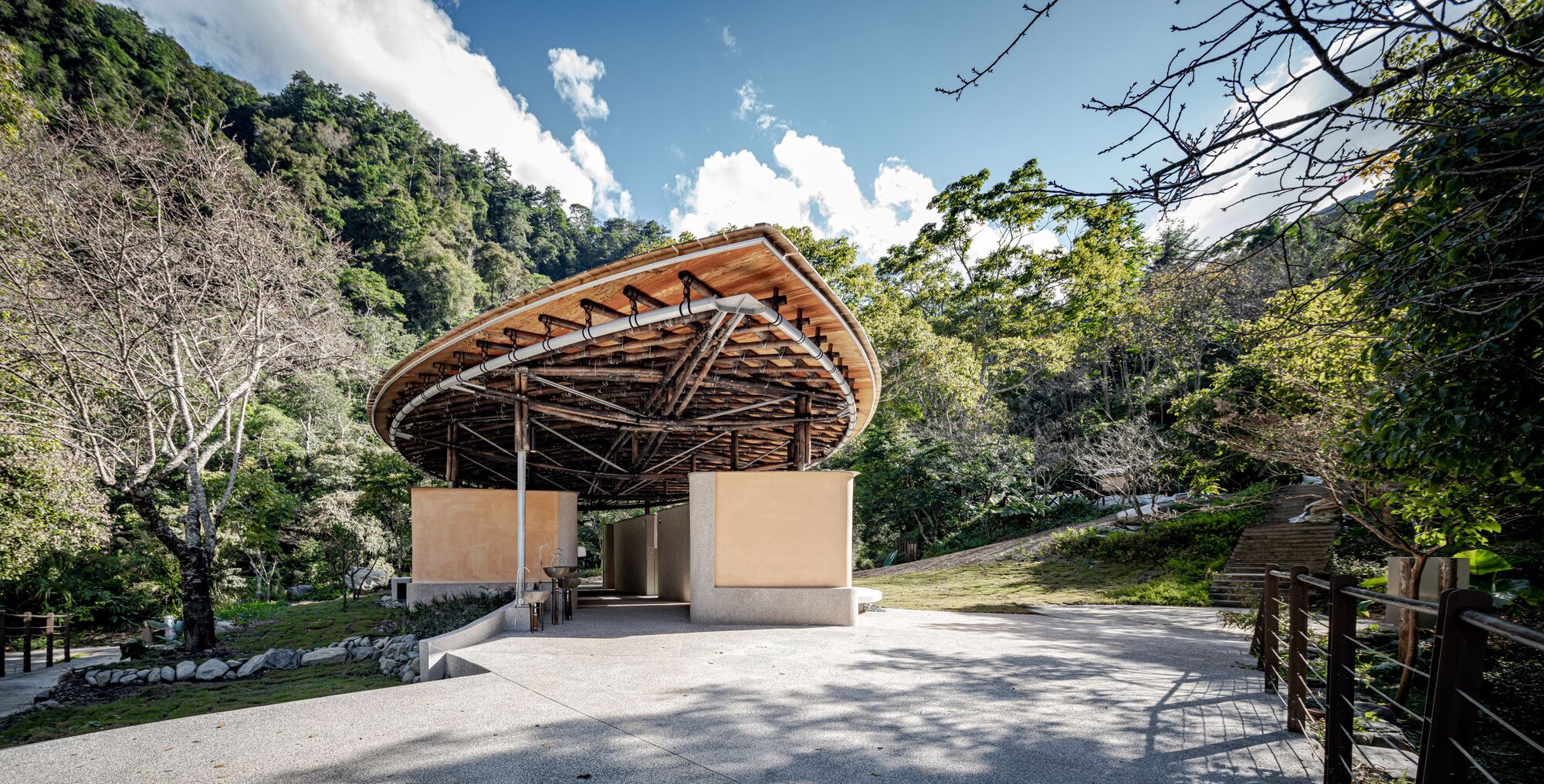 Wide view of the bamboo-ribbed canopy and ochre wall blocks on a paved terrace, with a timber railing and stepped path beside it