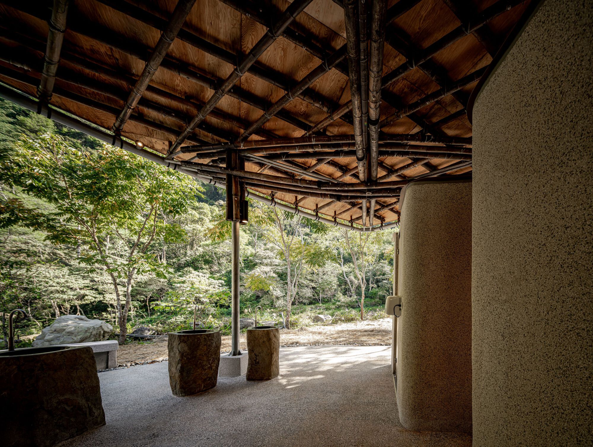 Open washing area under a curved timber roof with bamboo rafters, stone basins and a wooded hillside in dappled summer light