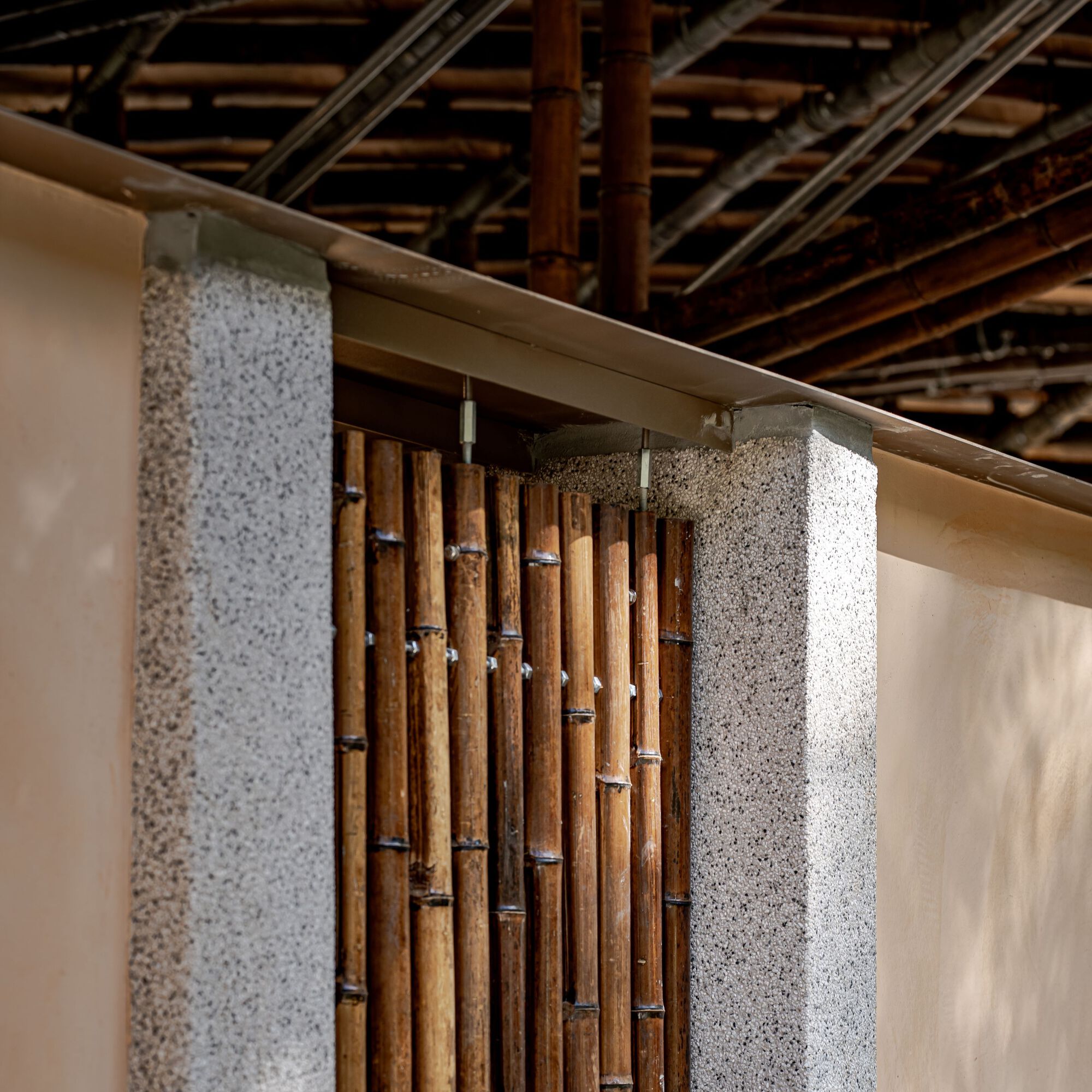 Close-up of vertical bamboo slats set between speckled stone piers beneath a bamboo-framed eave, with soft light on the plaster walls