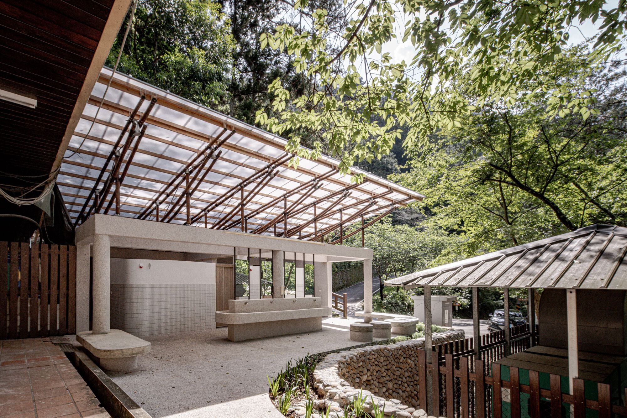 Courtyard with a translucent canopy on slender timber supports, stone benches and a low river-stone wall, framed by green summer trees