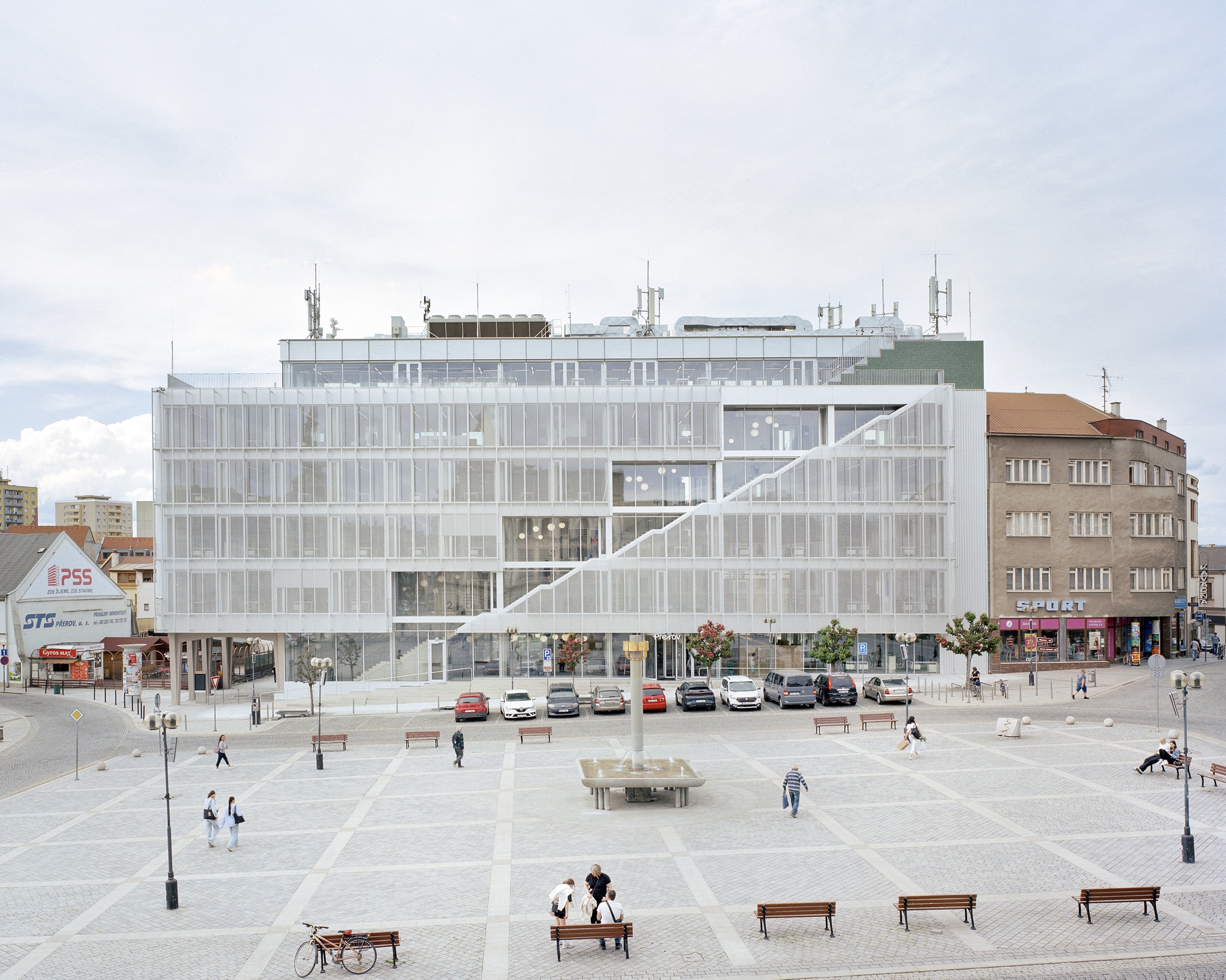 Translucent mesh-clad facade with a diagonal exterior stair above a paved square with benches, parked cars and pedestrians under overcast sky