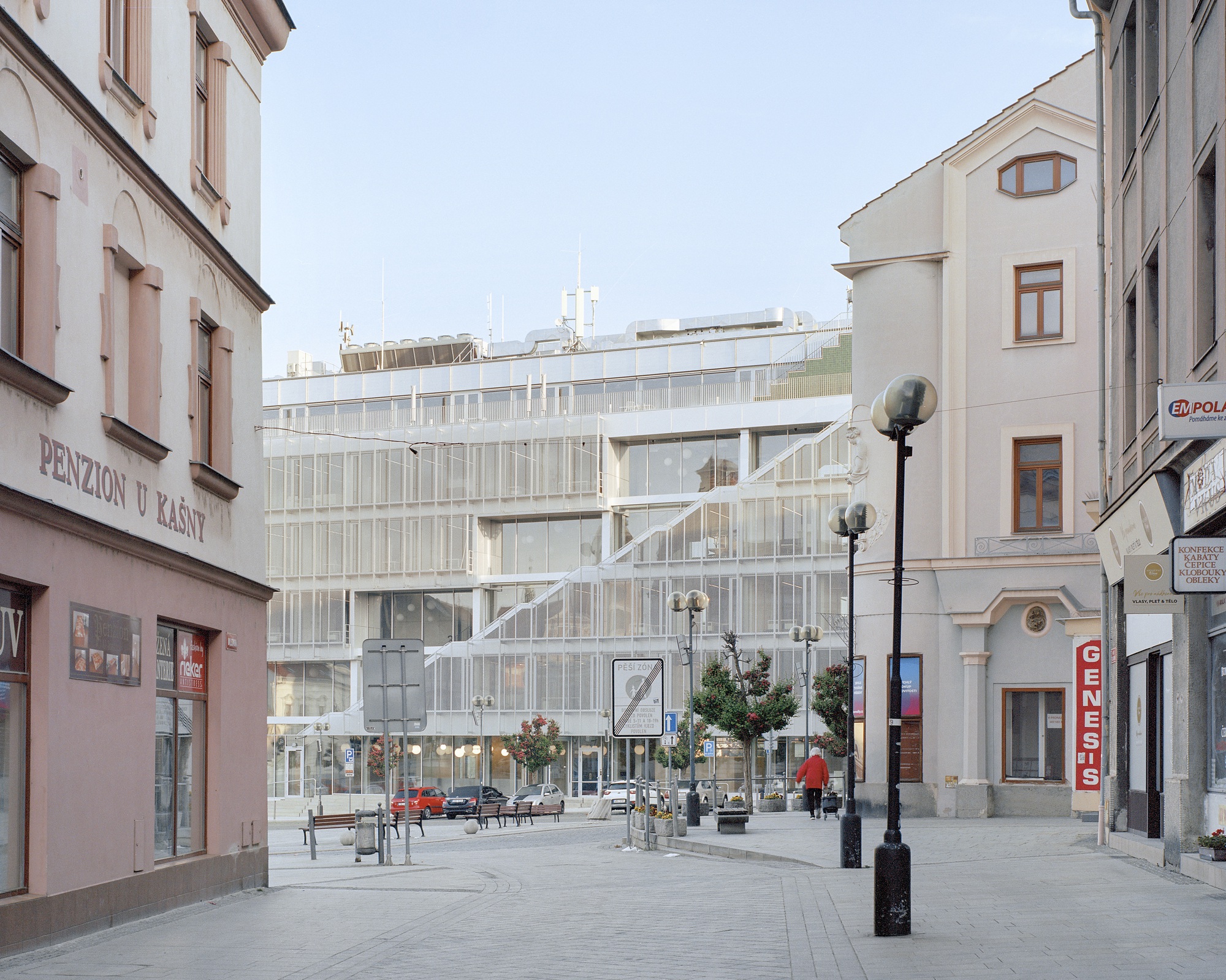Street view between older plastered buildings toward the glazed, mesh-wrapped block with its diagonal stair, a pedestrian in red passing below