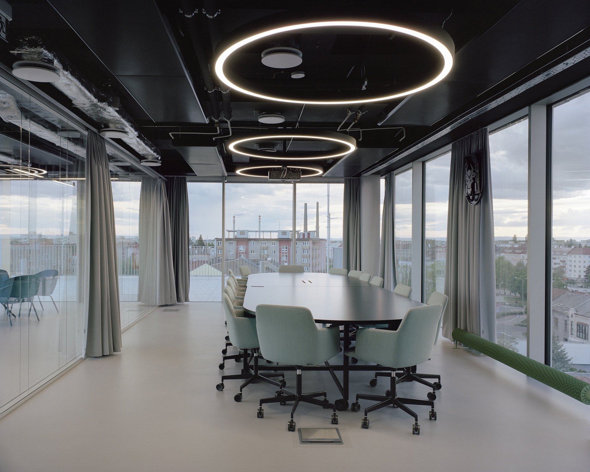 Conference room with an oval table, pale green chairs and three ring-shaped pendant lights beside tall windows with grey curtains in cloudy light