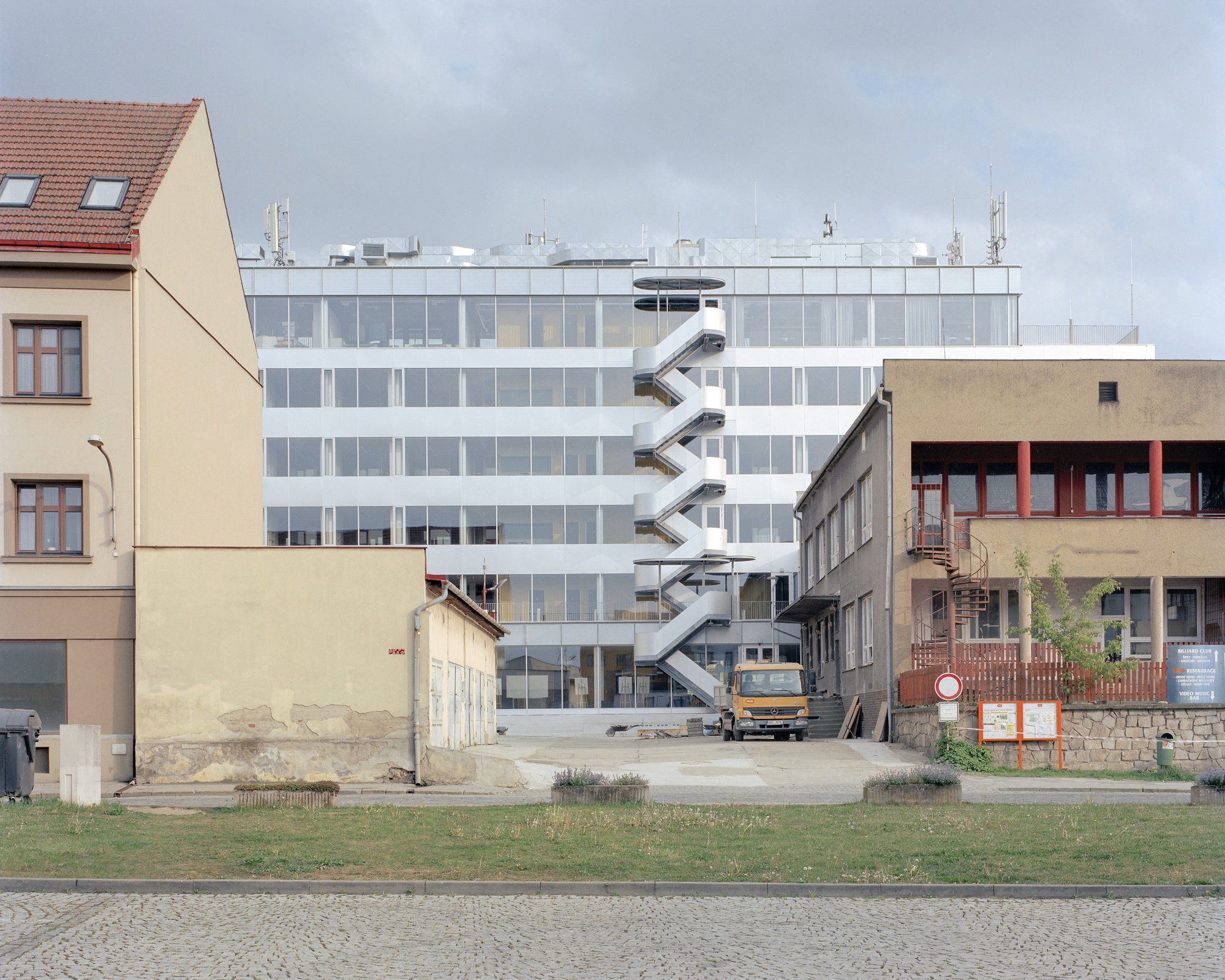 Rear view of the white glazed block with a zigzag steel fire stair, a yellow truck and low older buildings under grey cloud