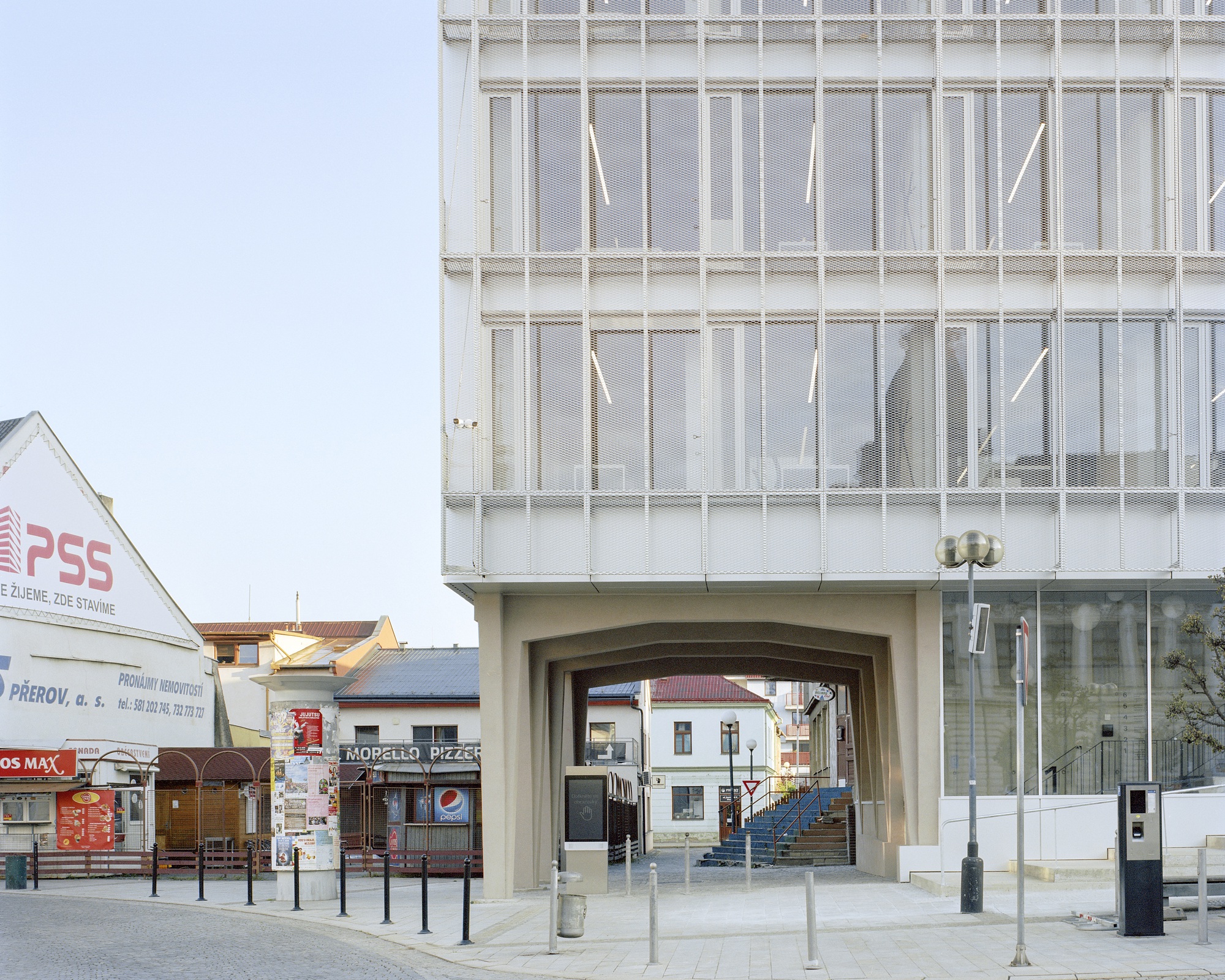 Concrete arched passage beneath a mesh-clad glass upper volume, with a stair beyond and a street lamp in clear morning light