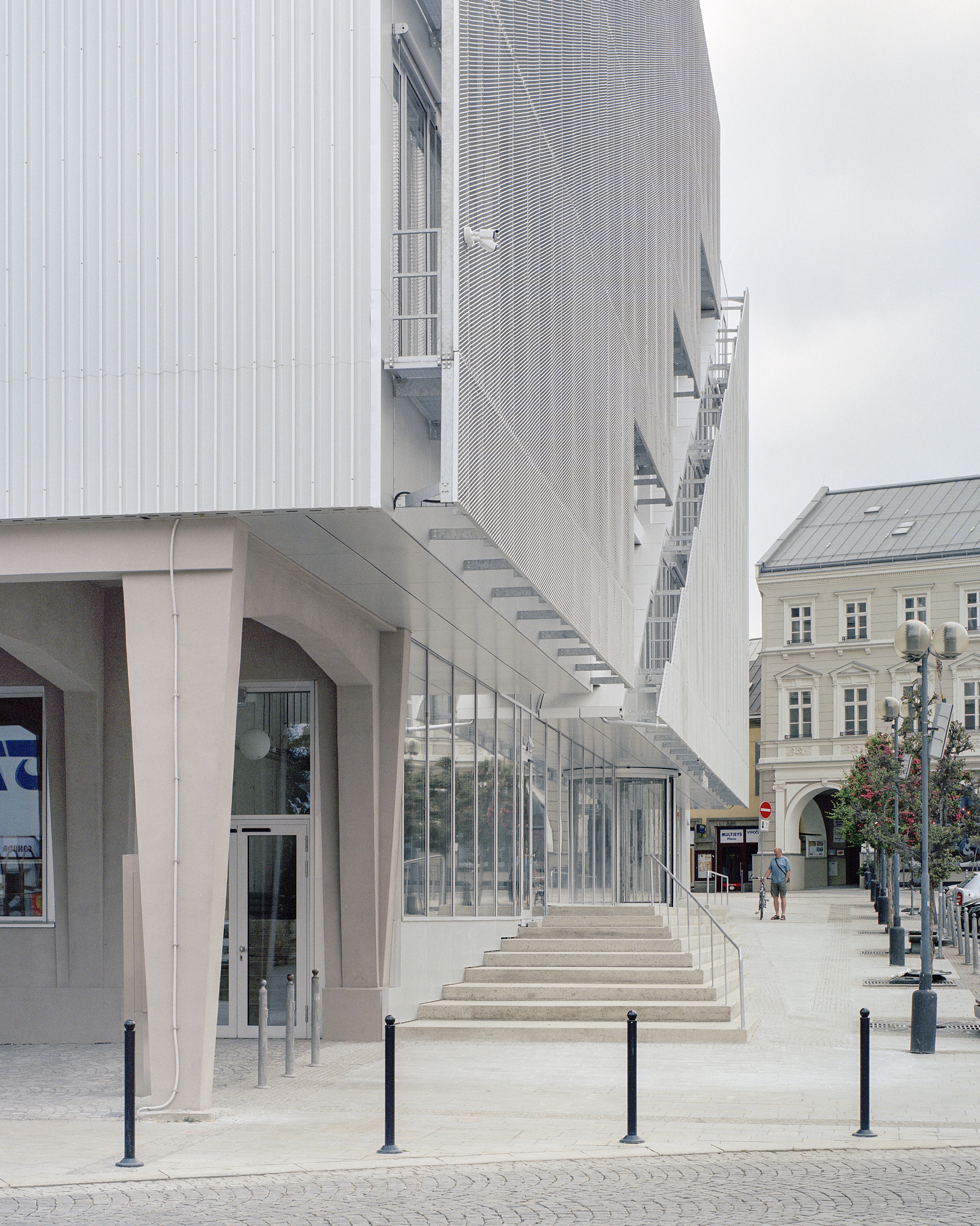 Tapered concrete columns and entrance steps below corrugated and perforated metal cladding, with a pedestrian walking in the square on an overcast day