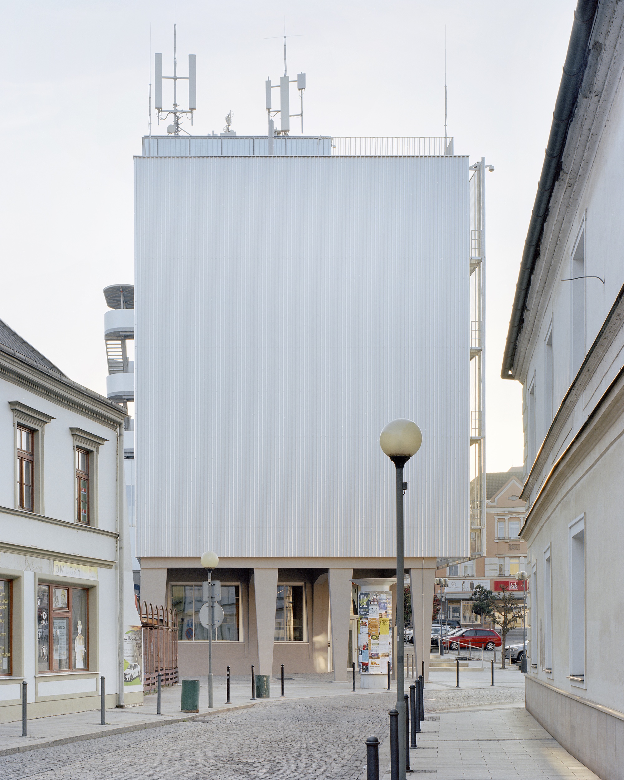 Blank corrugated metal tower block raised on concrete legs, topped with antennas, seen down a cobbled street at dusk
