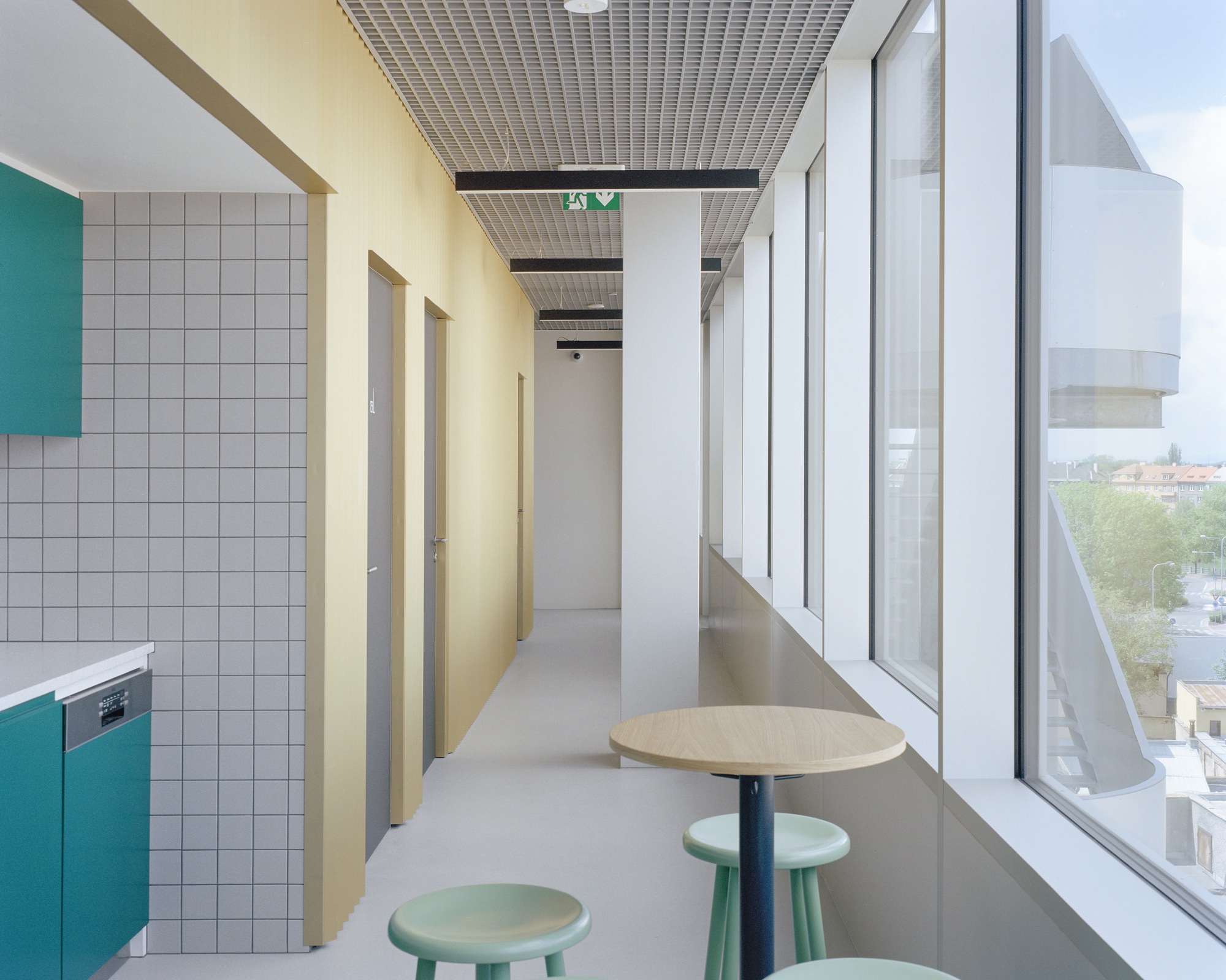 Narrow upper-floor corridor with yellow timber wall, teal kitchen cabinets, grey tile and round stools beside a long window wall in daylight