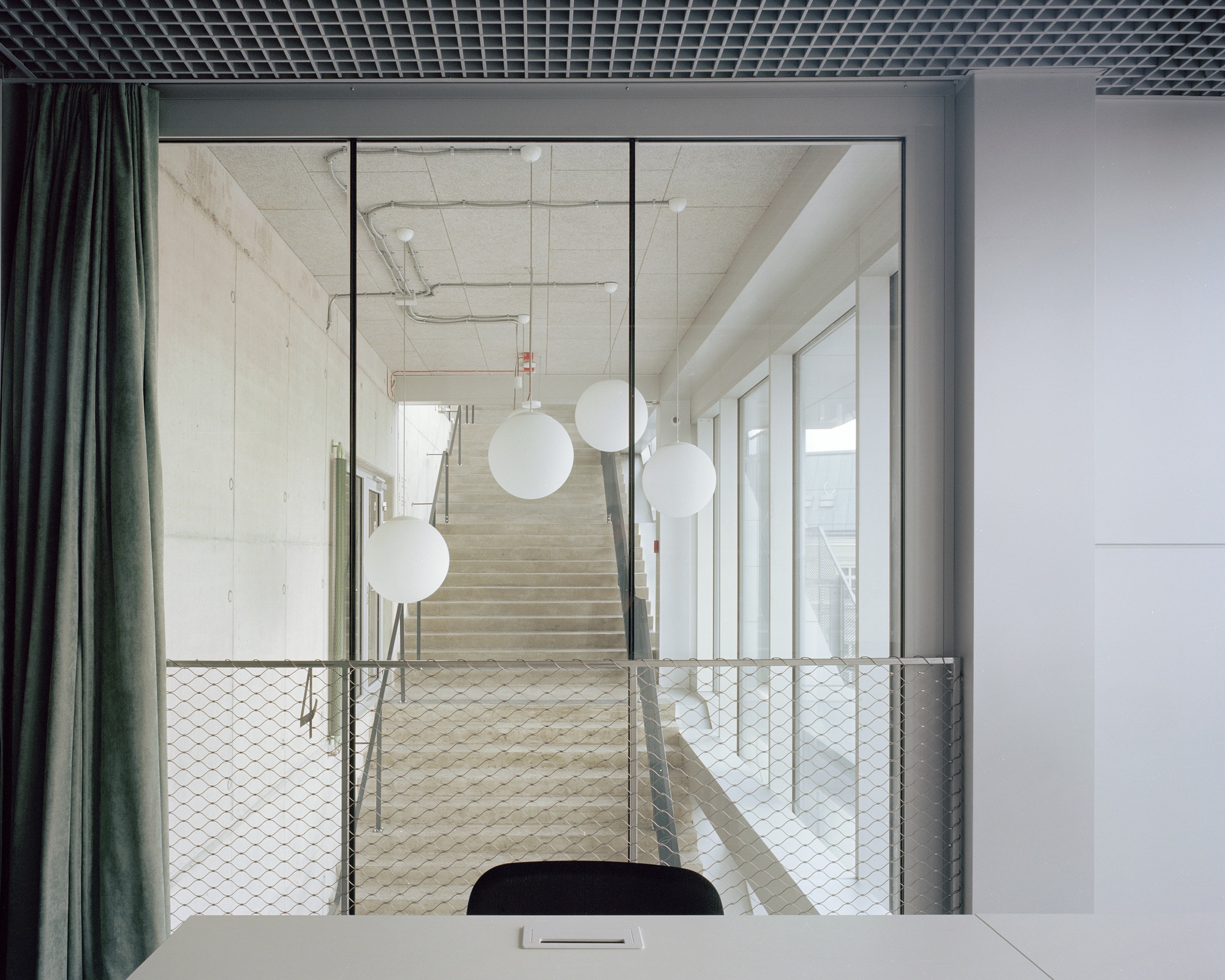 View from a desk through a glass partition to a concrete stair, with white globe pendant lamps and a green velvet curtain