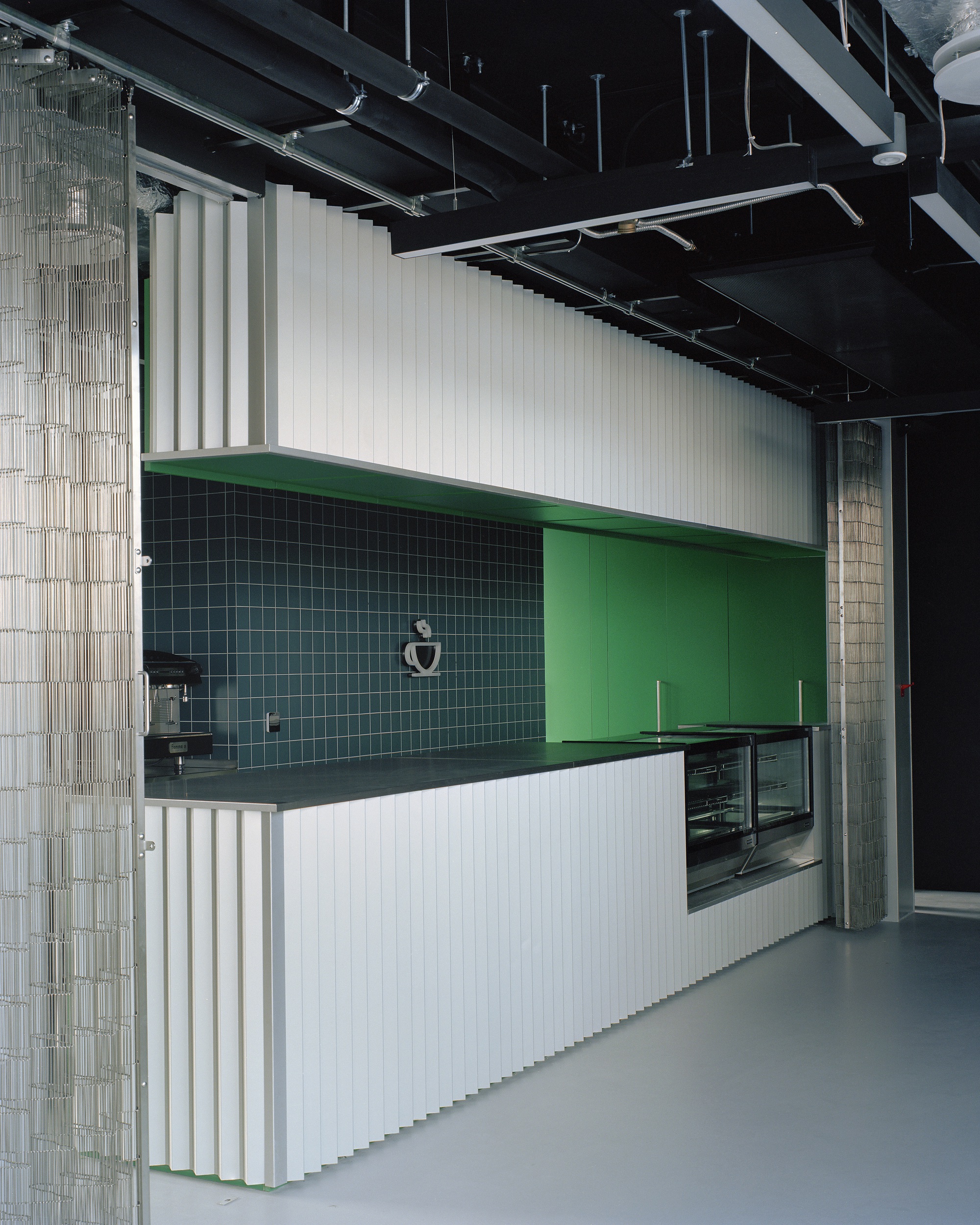 Pantry counter wrapped in white folded fins, with green cabinet doors, dark blue tile backsplash and a black exposed ceiling above