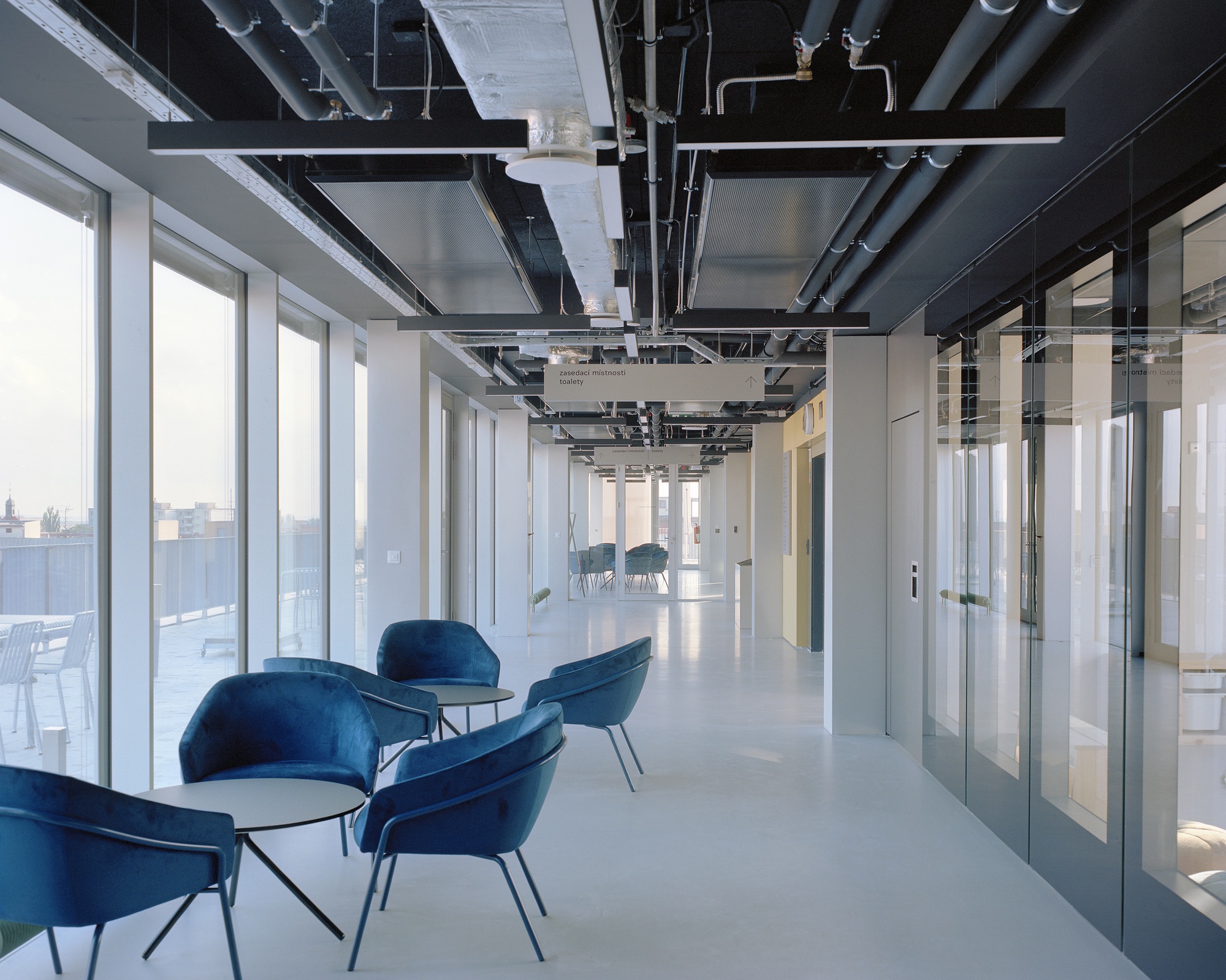 Top-floor corridor with blue velvet armchairs, exposed black ceiling pipes and a glazed partition wall at dusk