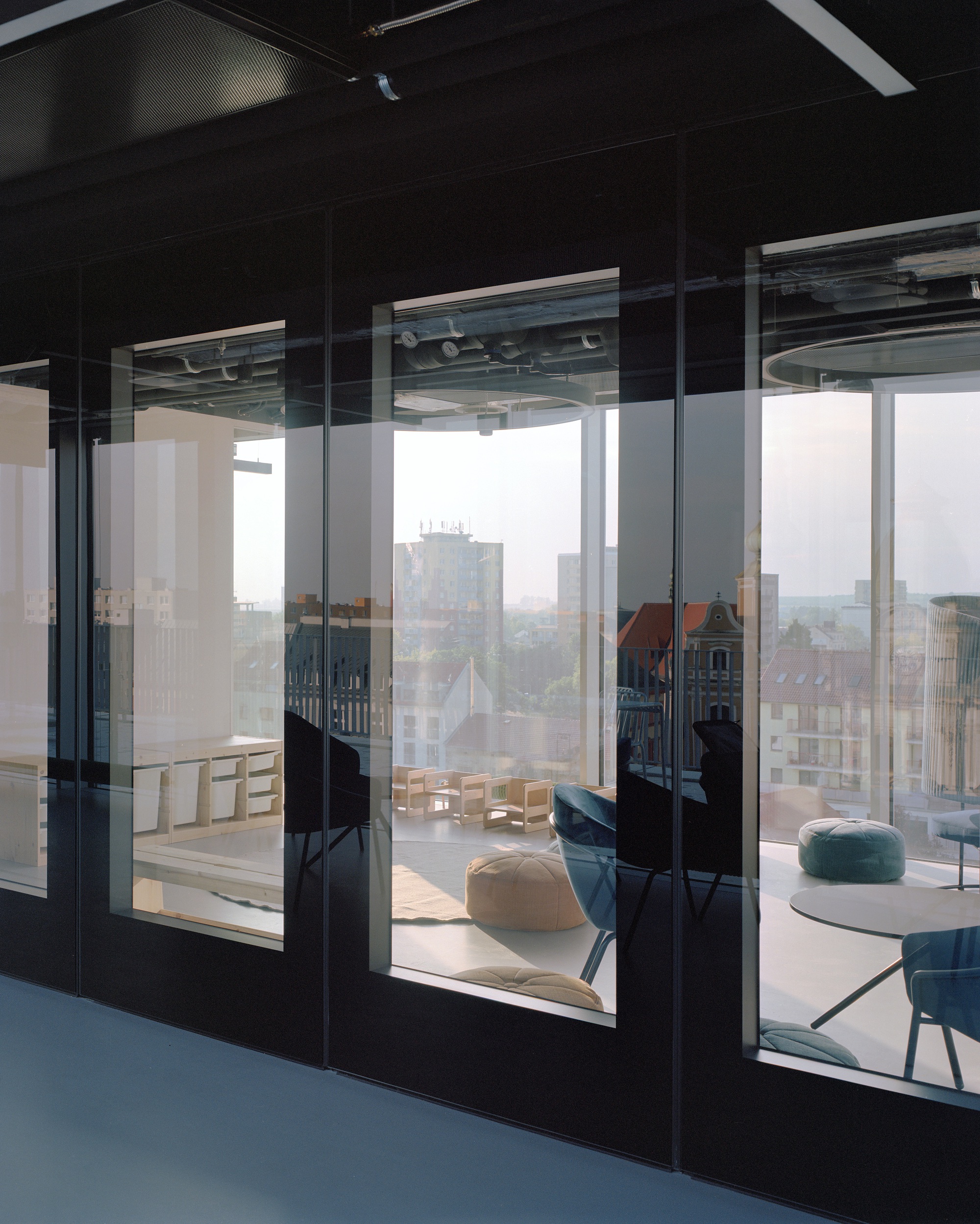 Dark framed glass partitions reflecting rooftops and a red-tiled gable, with a children's play room and floor cushions beyond