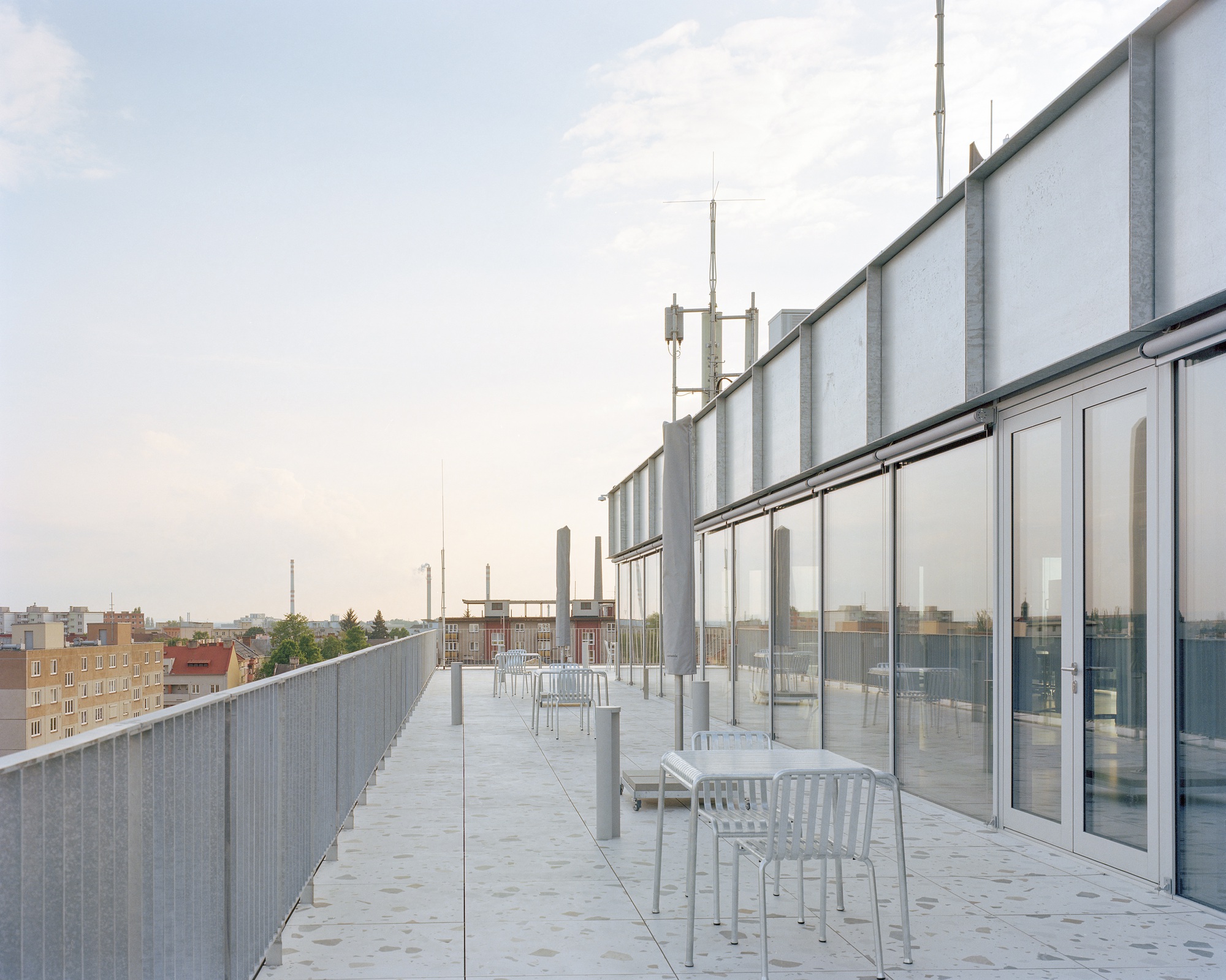 Rooftop terrace with terrazzo paving, metal chairs and a mesh railing facing low town buildings under a pale evening sky