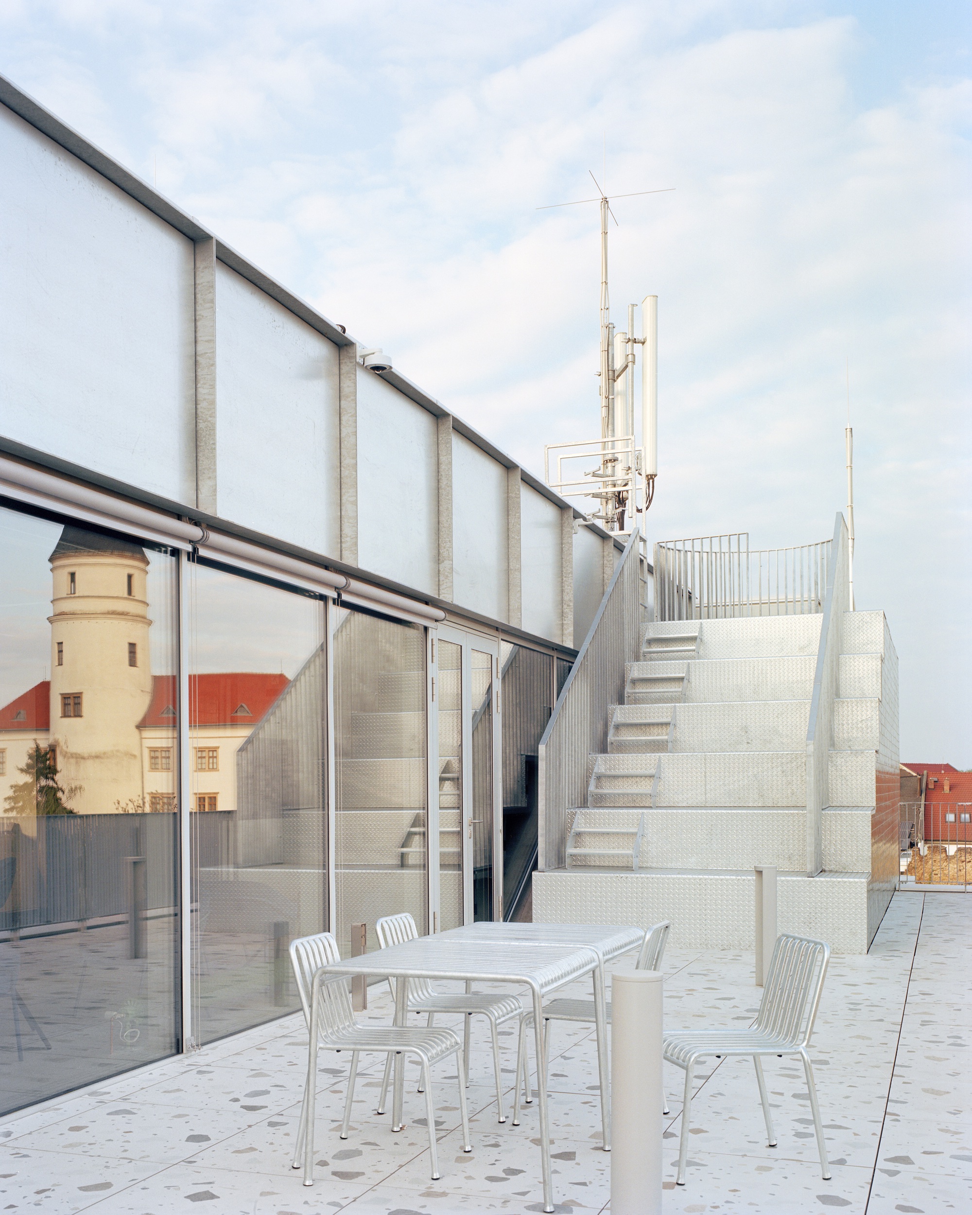 Metal staircase climbing to a rooftop antenna mast, with a church tower reflected in the glazing beside a terrace table