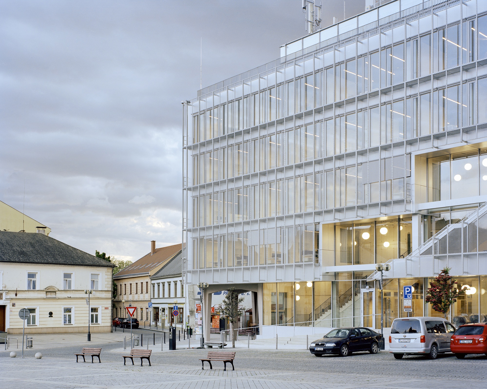 Glazed multi-storey facade with an open ground-floor arcade facing a town square with benches and parked cars under heavy grey clouds