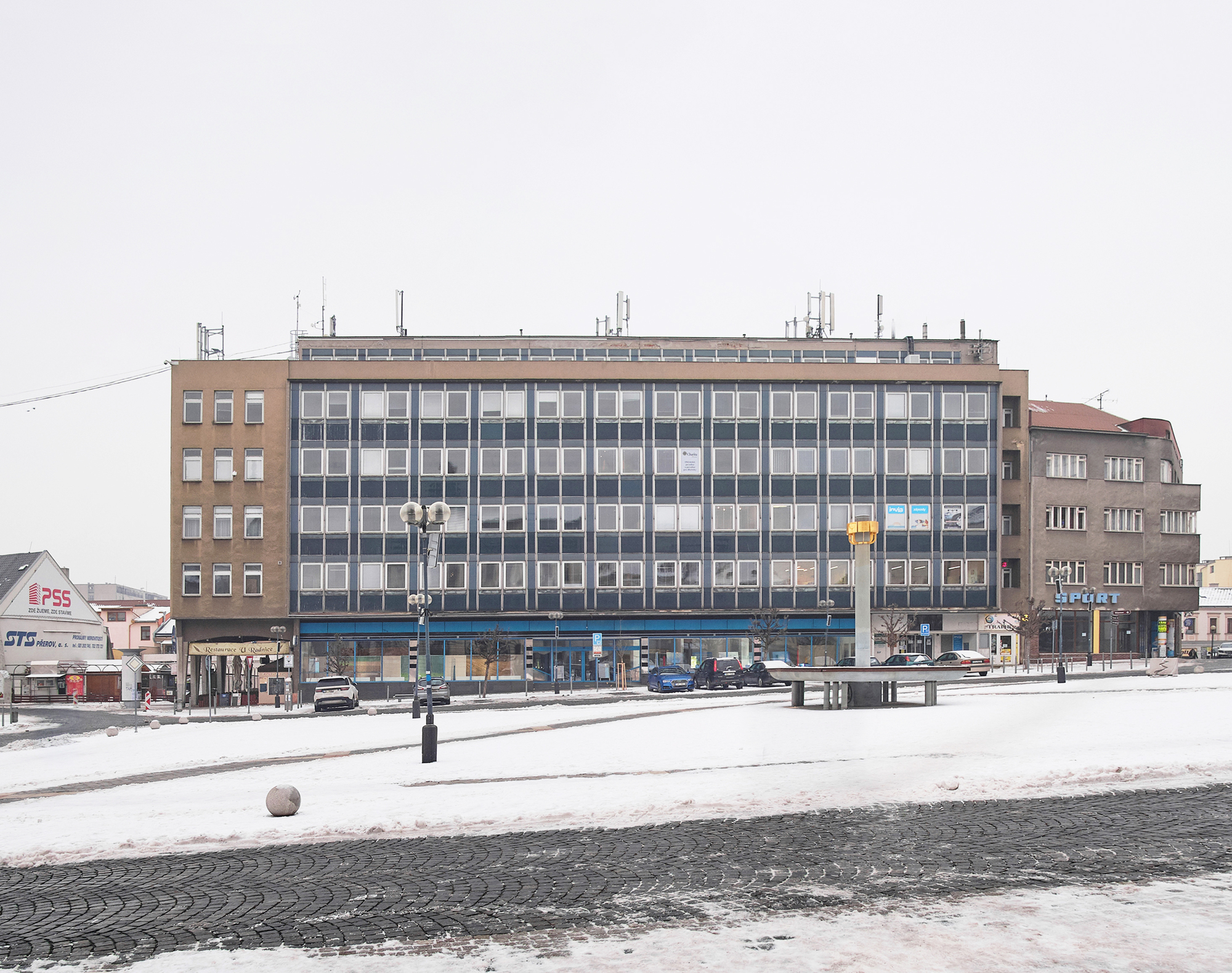 Five-storey office block with a blue-grey gridded window facade facing a snow-covered square under an overcast winter sky