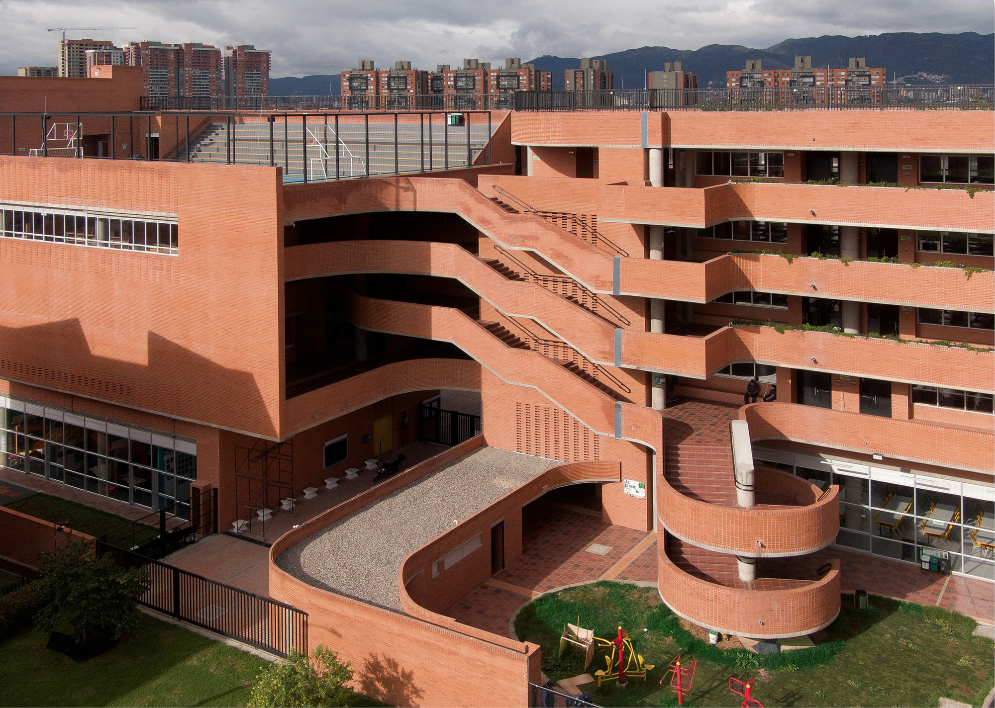 Curving brick stairs and terraced balconies stepping down to a courtyard with playground equipment under a cloudy sky
