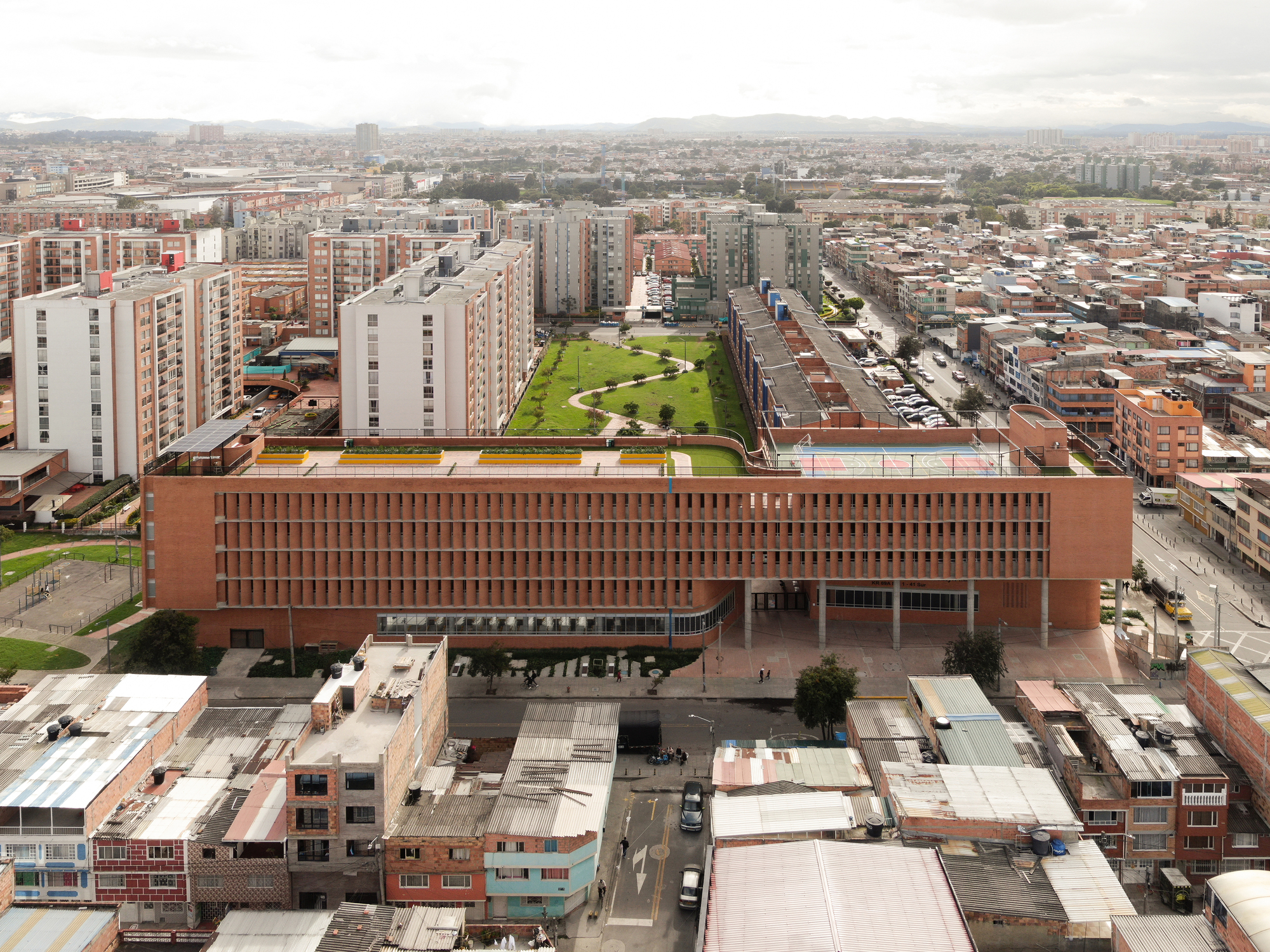 Aerial view of the long brick building with a perforated facade and green roof garden above surrounding low-rise streets