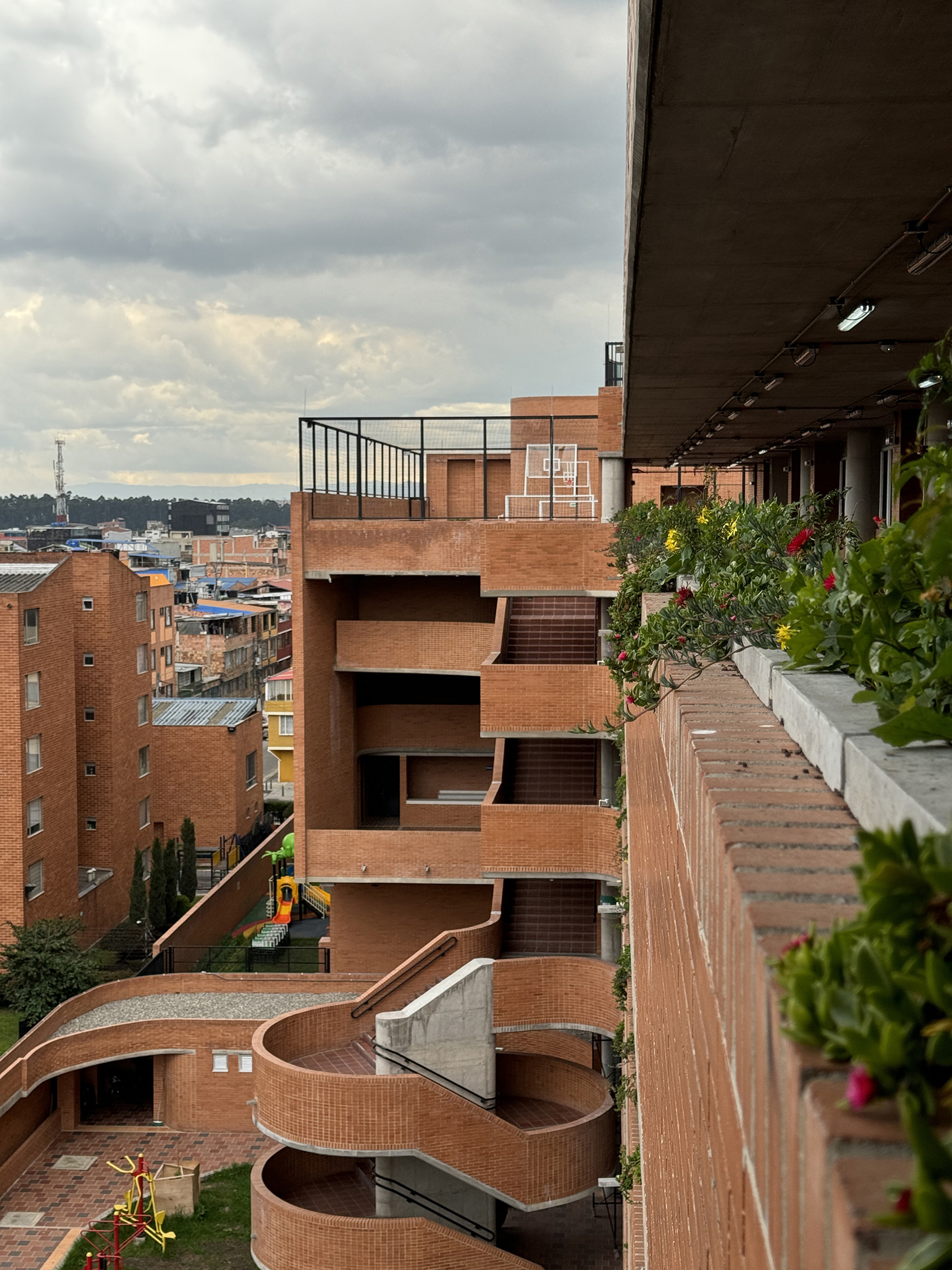 Brick spiral stair and stepped terraces seen past a planter of red and yellow flowers on an overcast day