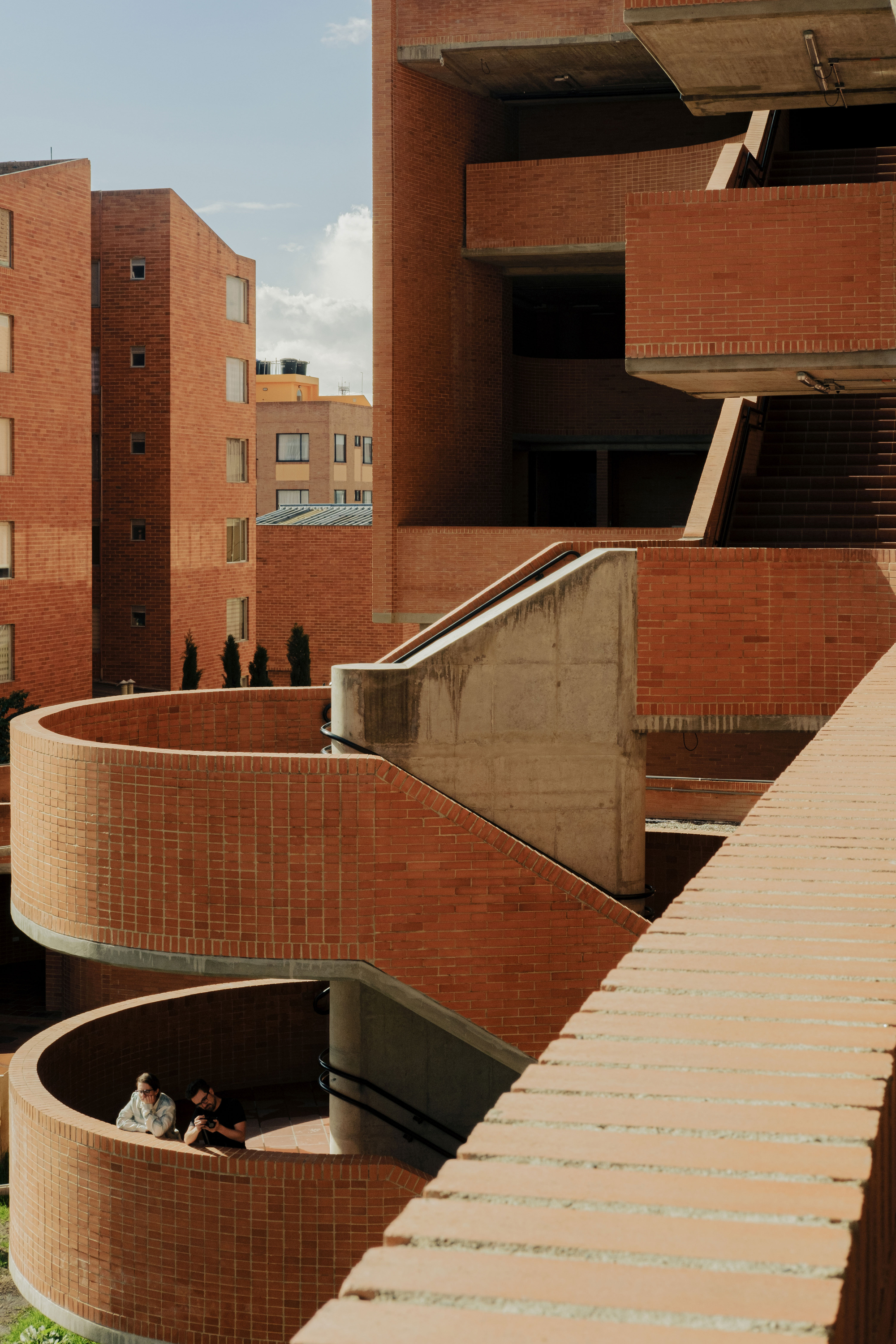Curved brick stair landing with a concrete wall, where two people lean on the rail in afternoon sun
