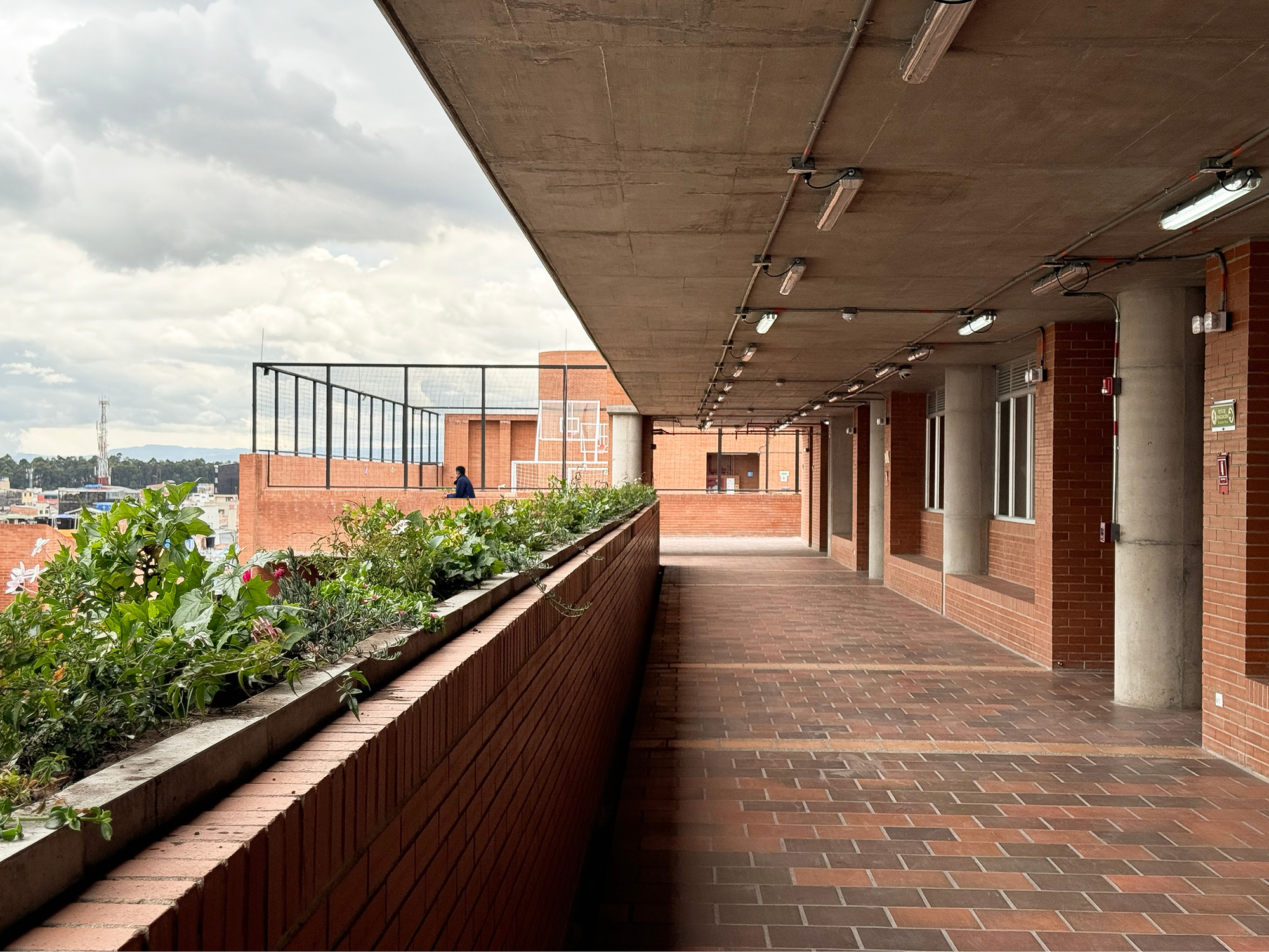 Open brick-paved gallery with concrete columns and a long planter bed, lit by tube lights under a concrete ceiling