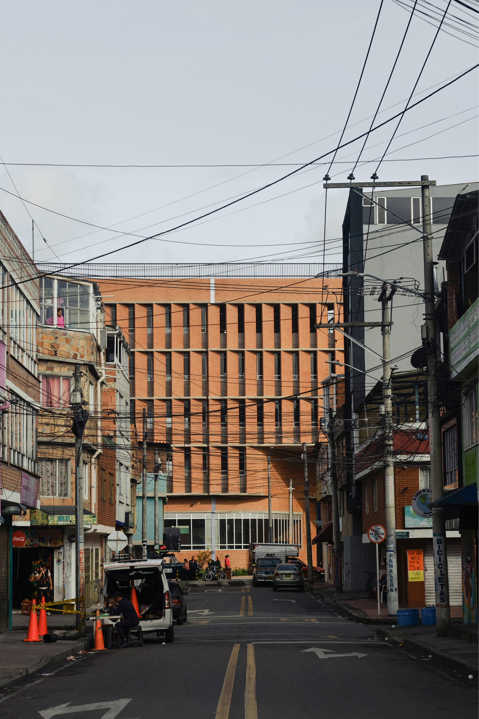 Brick block with vertical slot openings rising above a narrow street of low shops, utility poles and parked vans under an overcast sky