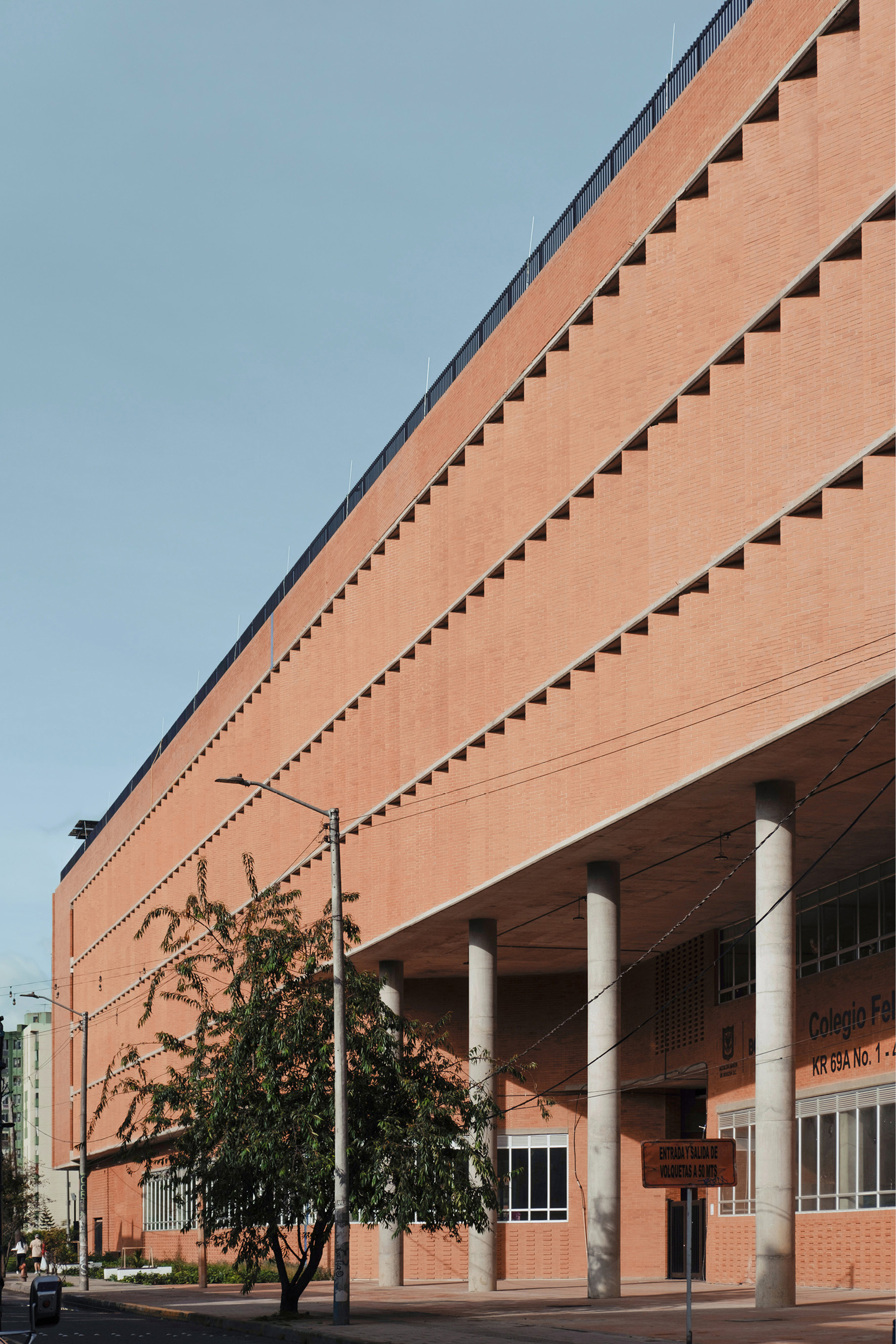 Long brick facade with stepped diagonal courses raised on slender concrete columns, a leafy tree in the foreground in clear morning light