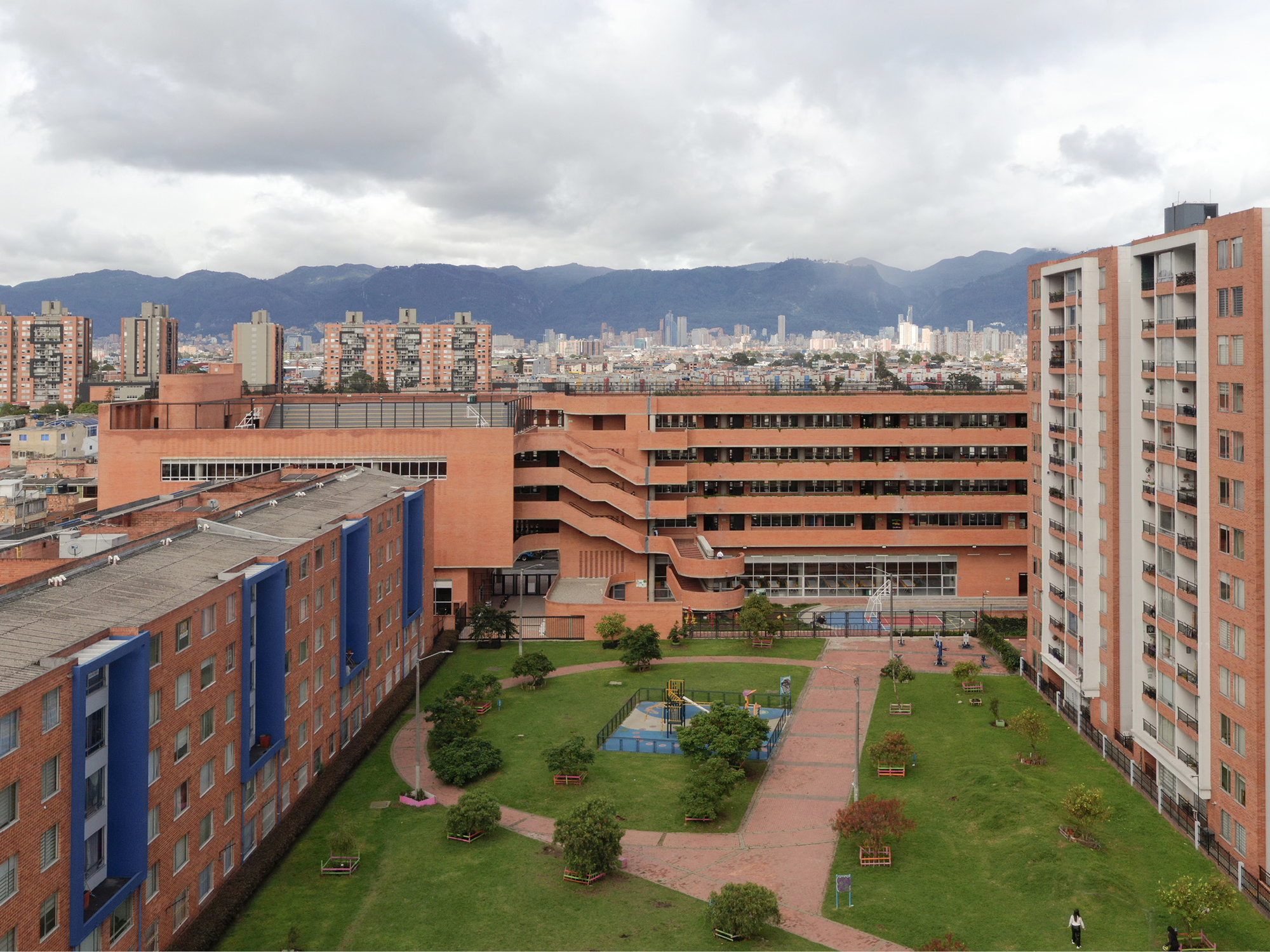 Elevated view of a landscaped courtyard with a curving brick path, playground and terraced brick school building beneath mountains and heavy clouds