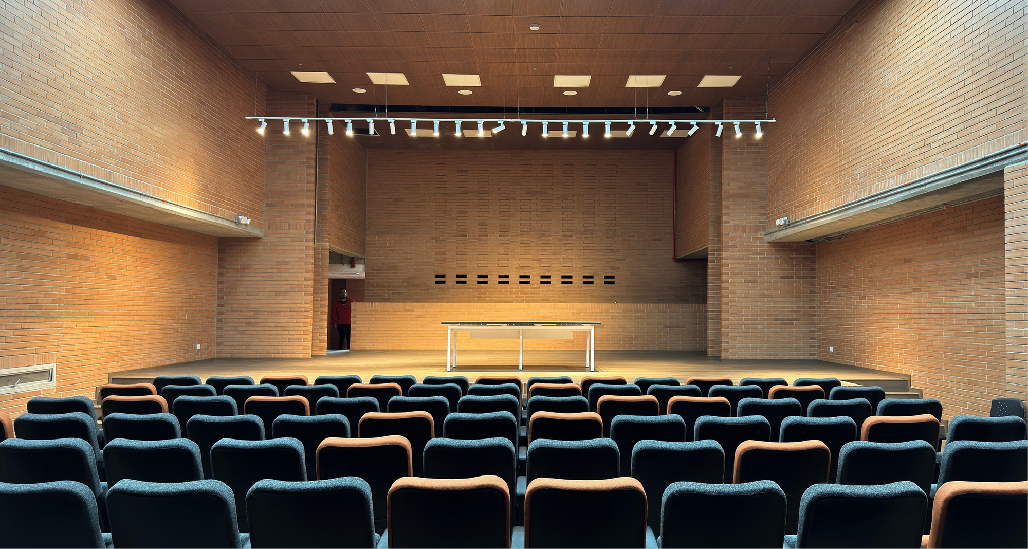 Brick-walled auditorium with rows of dark grey and orange seats facing a low stage, lit by a ceiling track of spotlights