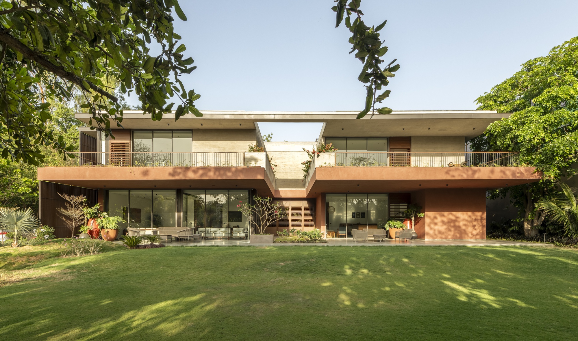 Two-storey garden facade with terracotta-toned cantilevered balconies and a central gap, seen across a lawn dappled by tree shadows