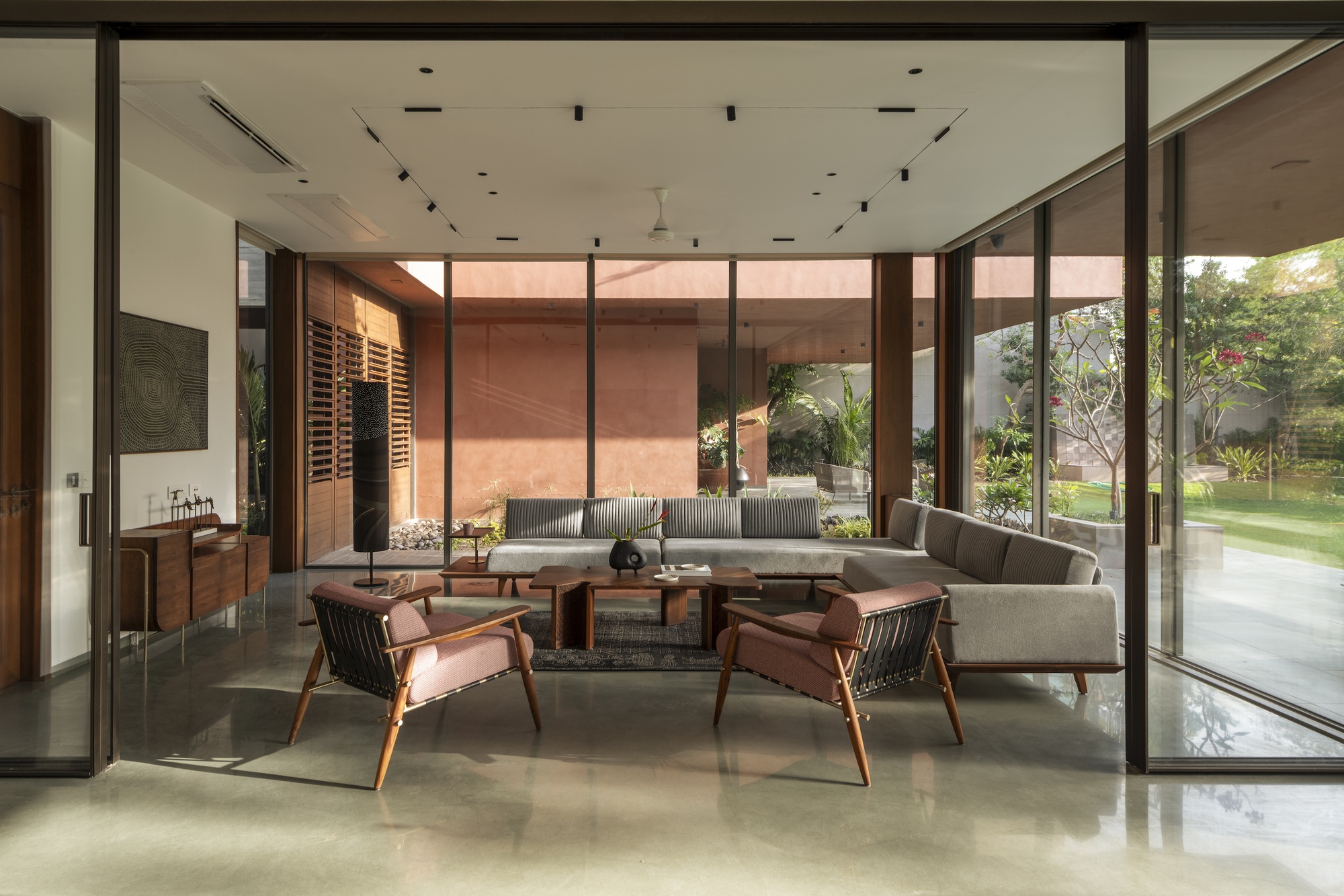 Living room with grey sofa, pink-cushioned timber armchairs and polished concrete floor opening through sliding glass to a sunlit lawn