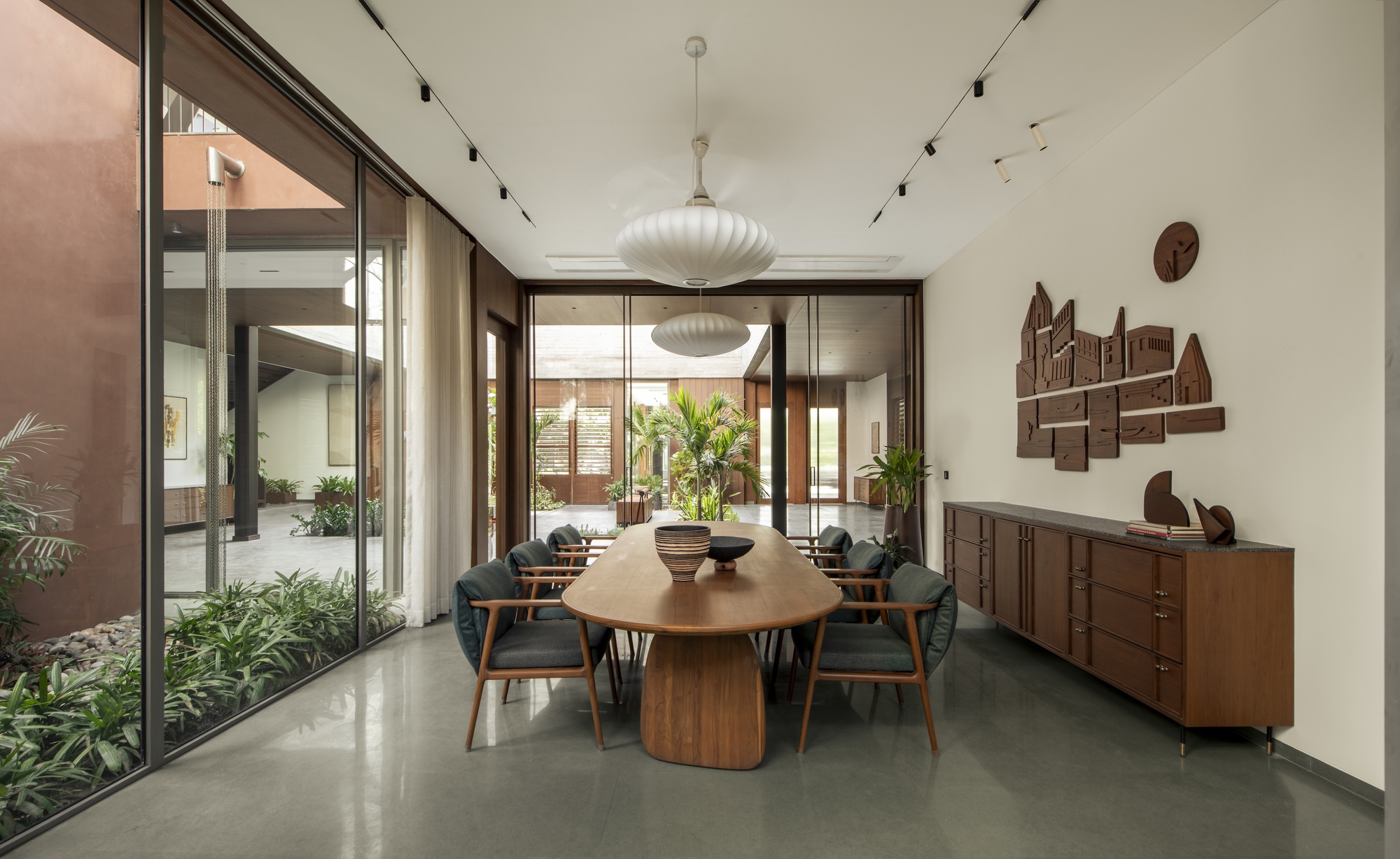 Dining room with an oval timber table, pendant lamps and a carved wall relief above a sideboard, glazed toward a courtyard garden
