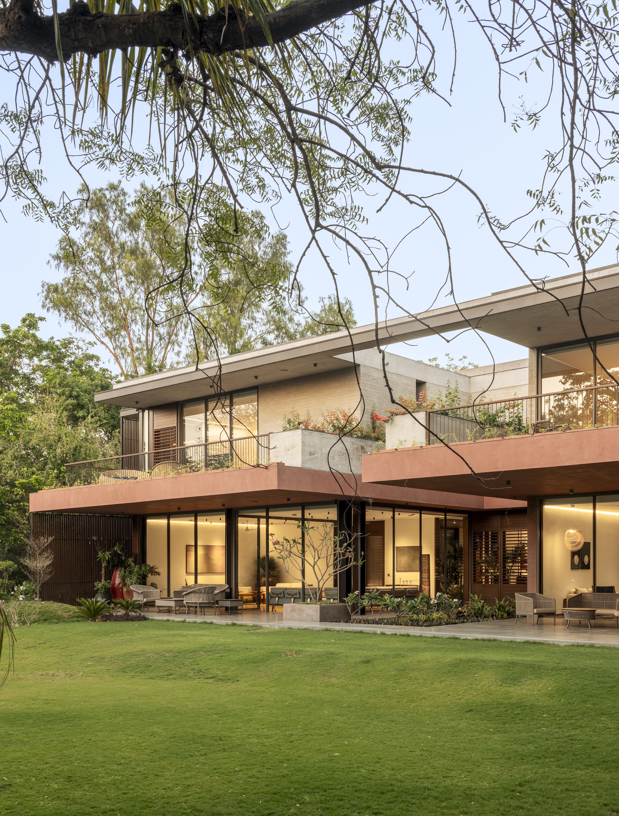 Two-storey house with terracotta-toned cantilevered slabs and glazed ground floor, seen across a lawn under hanging branches at dusk