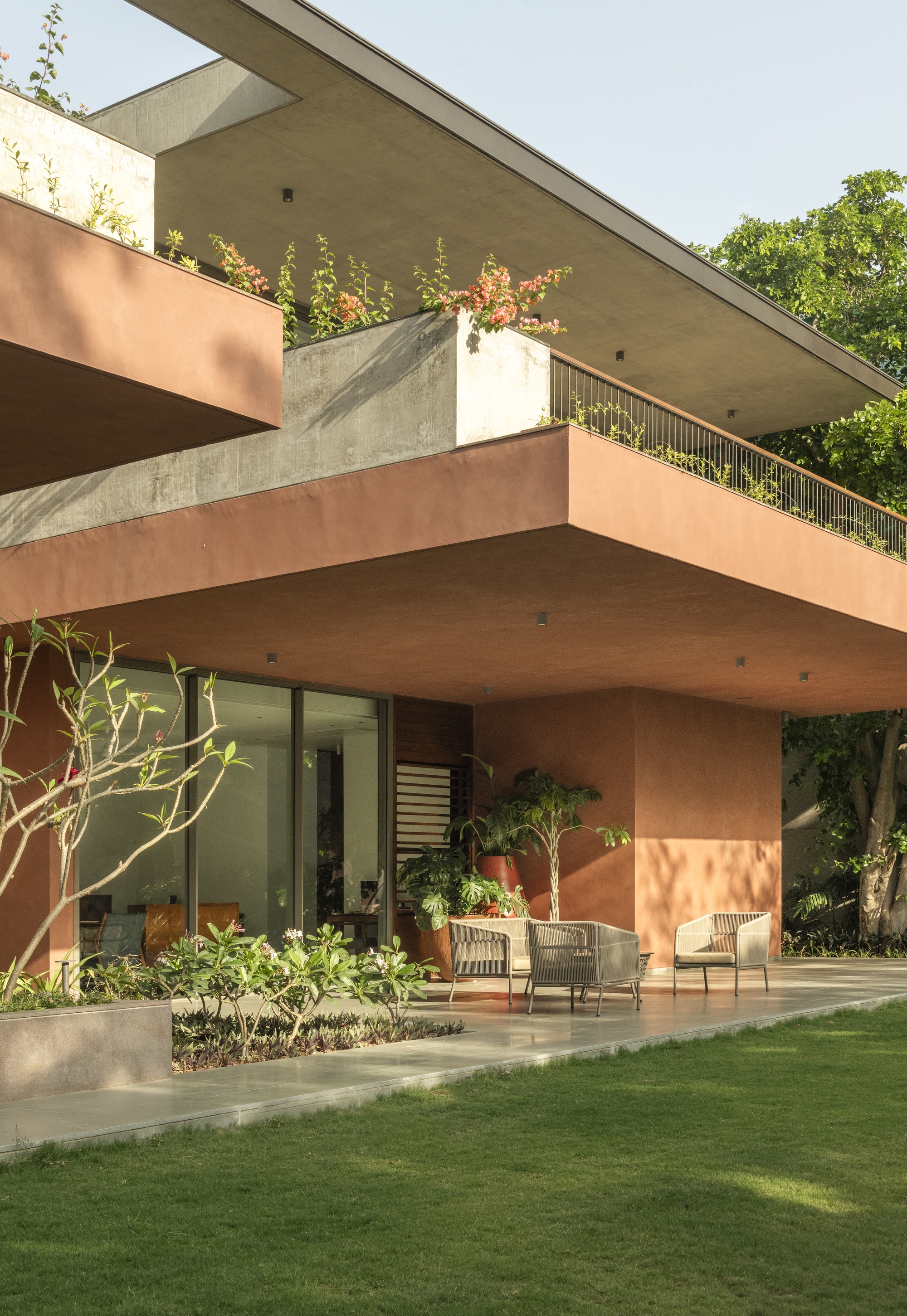 Terrace with wicker chairs under a deep terracotta overhang, concrete planter with flowering bougainvillea above, lawn in warm afternoon light